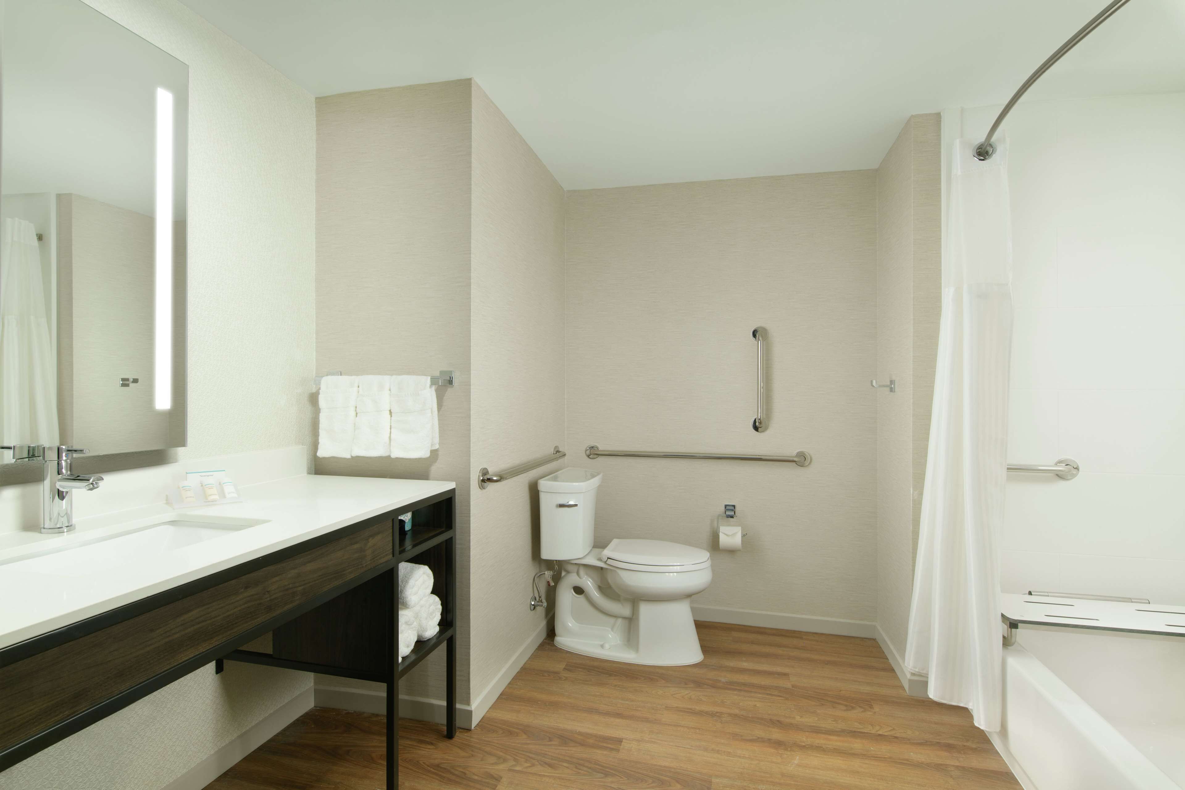 Un baño moderno con lavamanos, espejo y retrete blancos. Incluye zona de ducha con puerta de cristal y barra de apoyo.