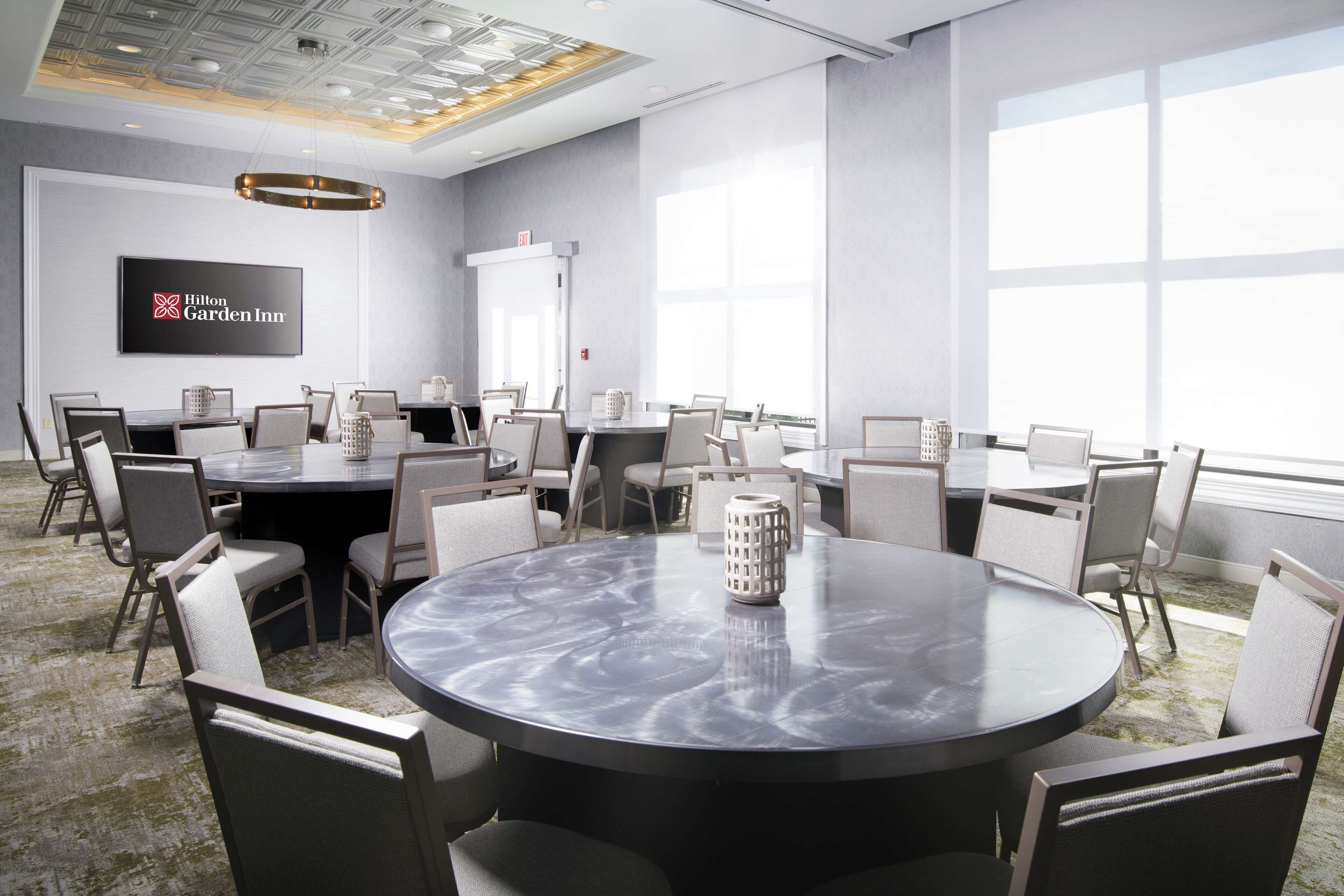 A conference room with round tables and chairs, a large window, and a Hilton Garden Inn sign on the wall.