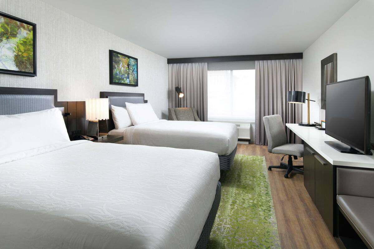 A hotel room with two beds, a desk, a chair, a TV, and a window with curtains.