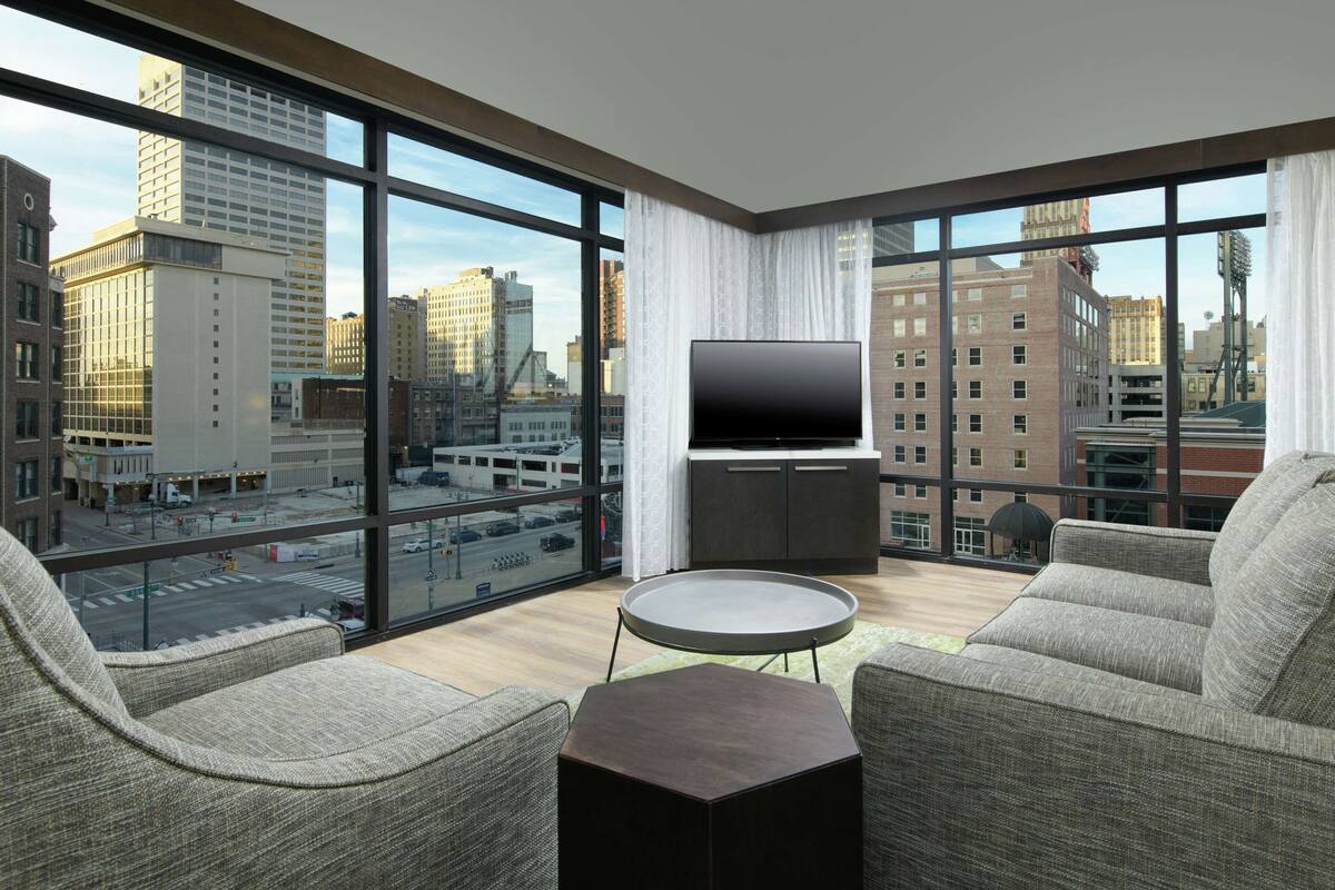 A modern hotel room with large windows, a flat-screen TV, and comfortable seating.