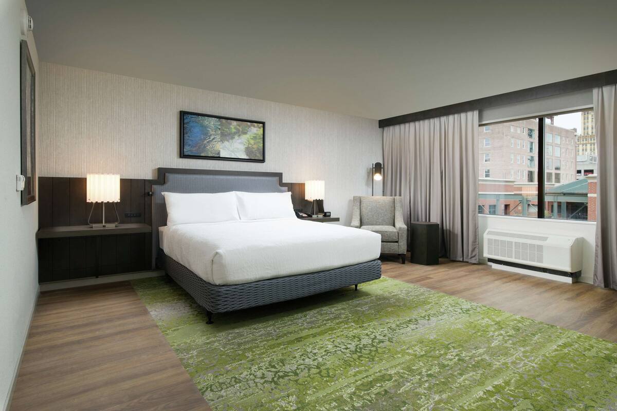 A hotel room with a large bed, a green patterned rug, a view of buildings through the window, and a painting on the wall.