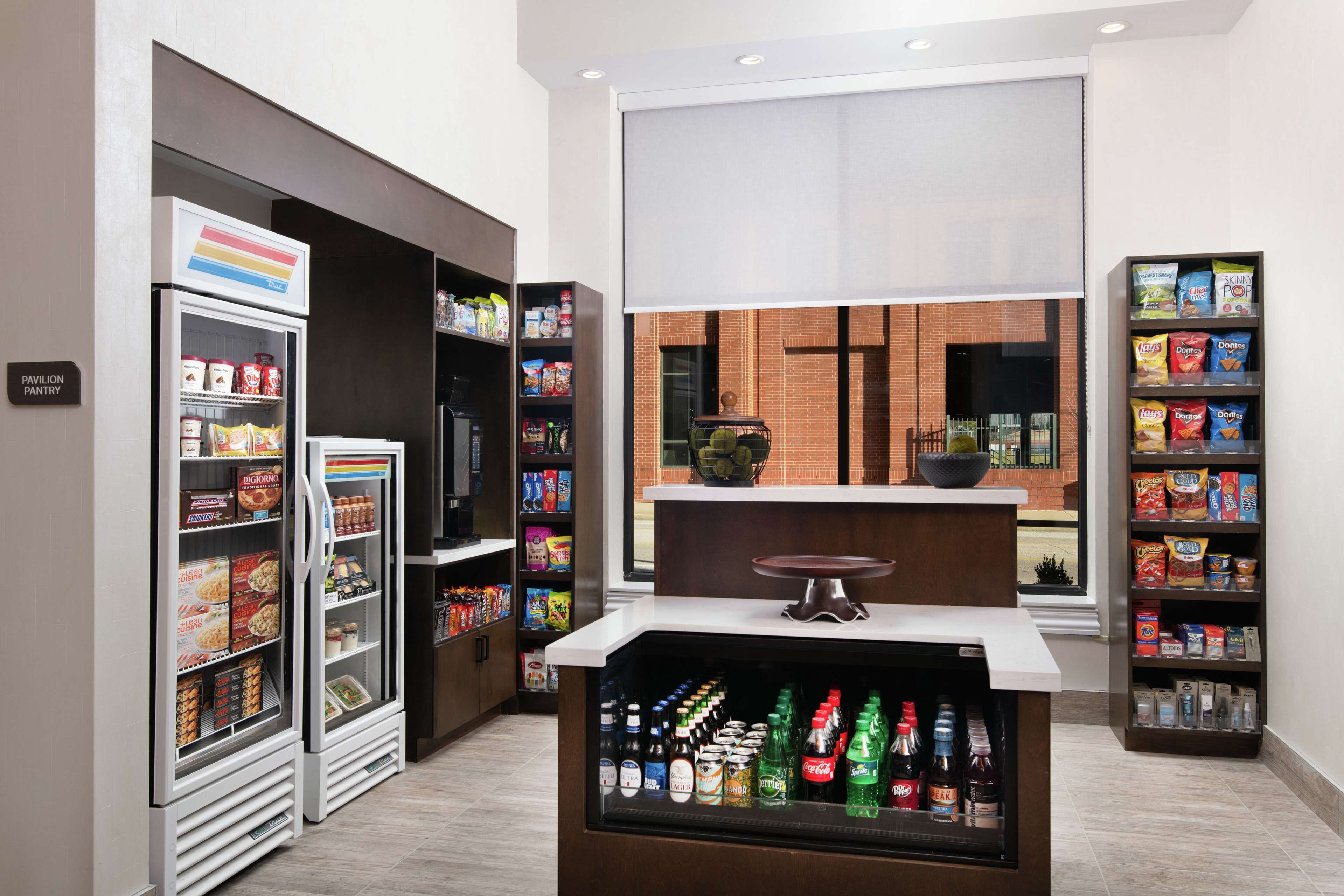 Uma despensa moderna de hotel com prateleiras para lanches refrigerados e não refrigerados, um refrigerador de bebidas e uma área de estar com vista.