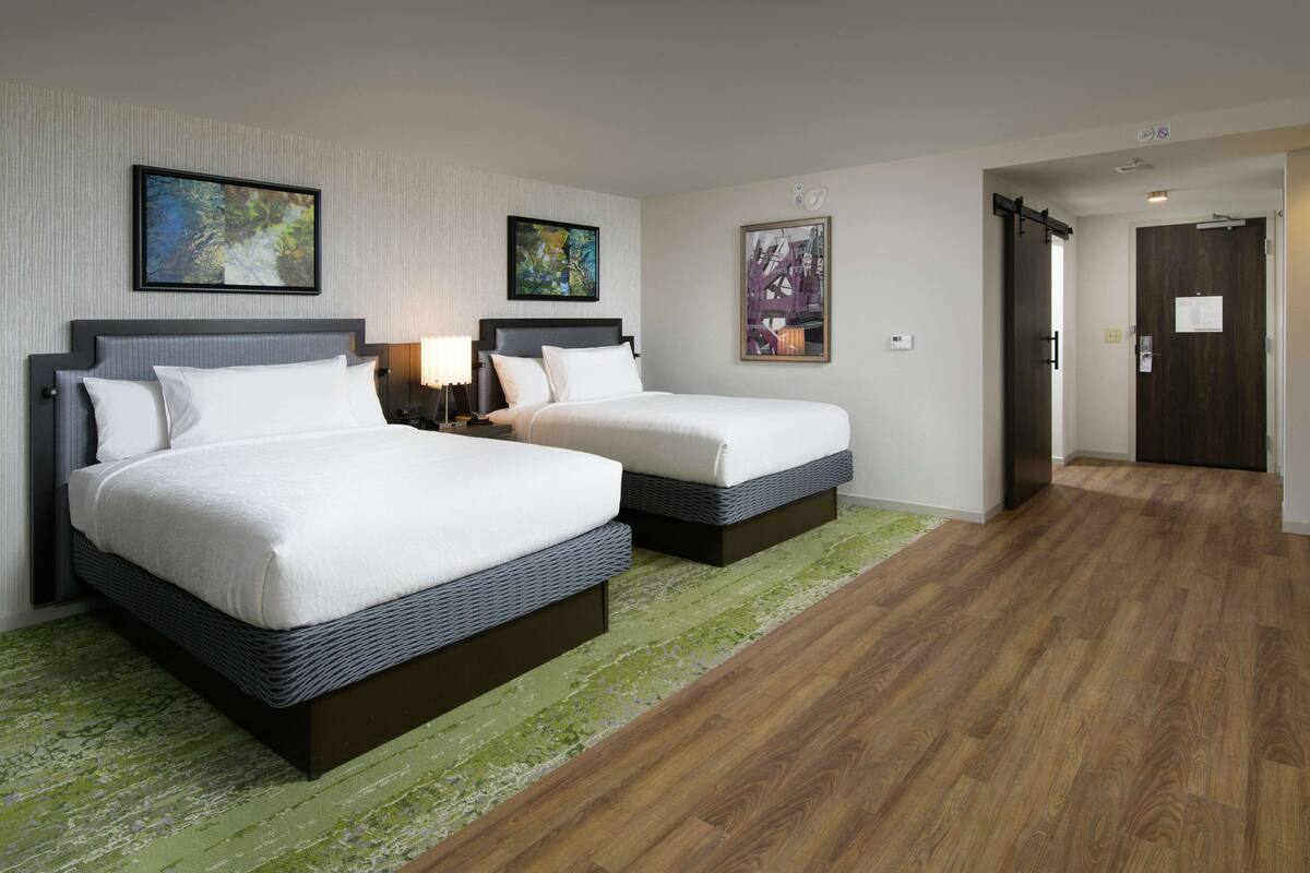 A hotel room with two beds, a green carpet, wooden flooring, and two framed pictures on the walls.