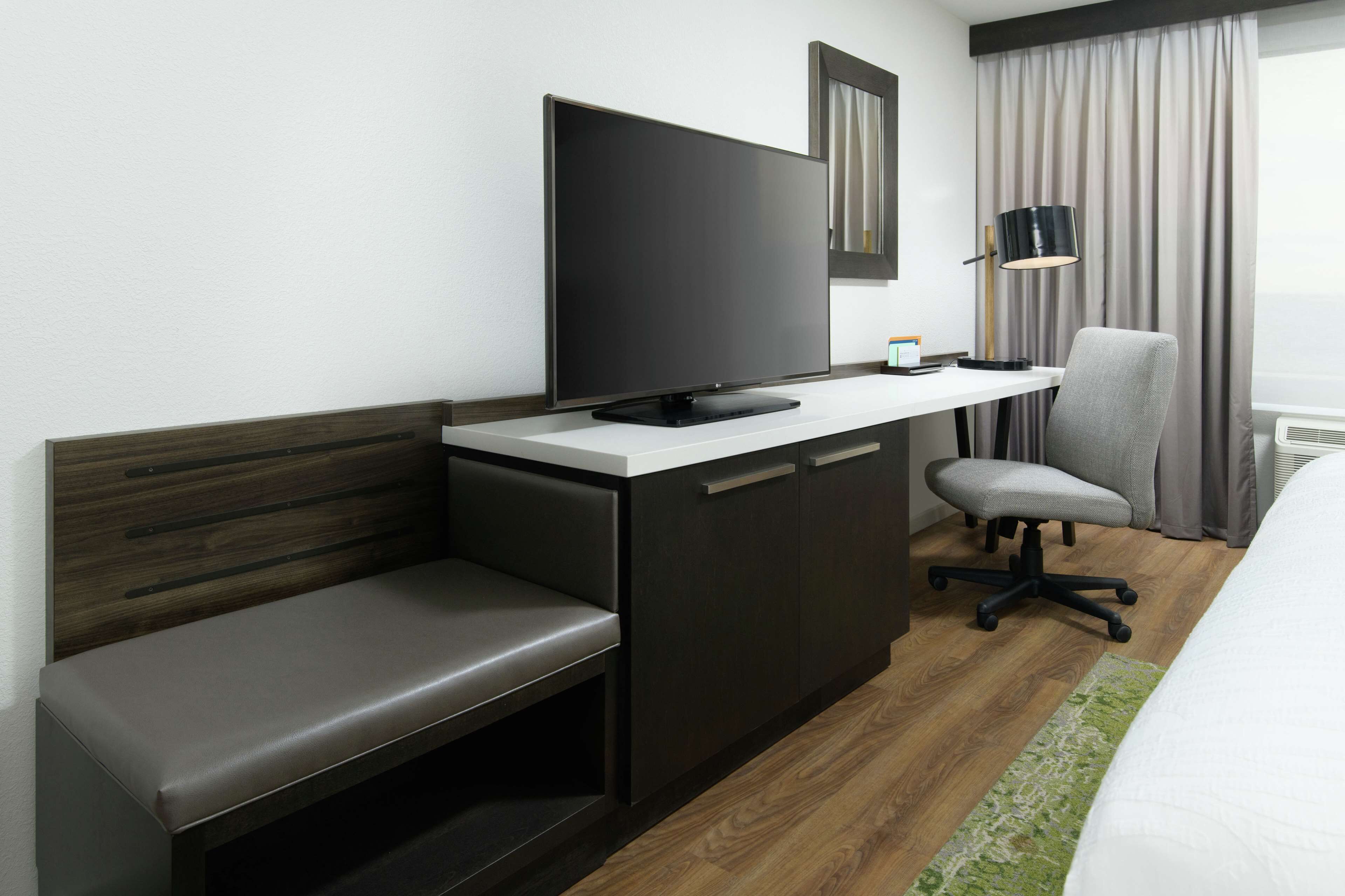 A hotel room with a bed, a desk with a flat-screen TV, a chair, and a bench.