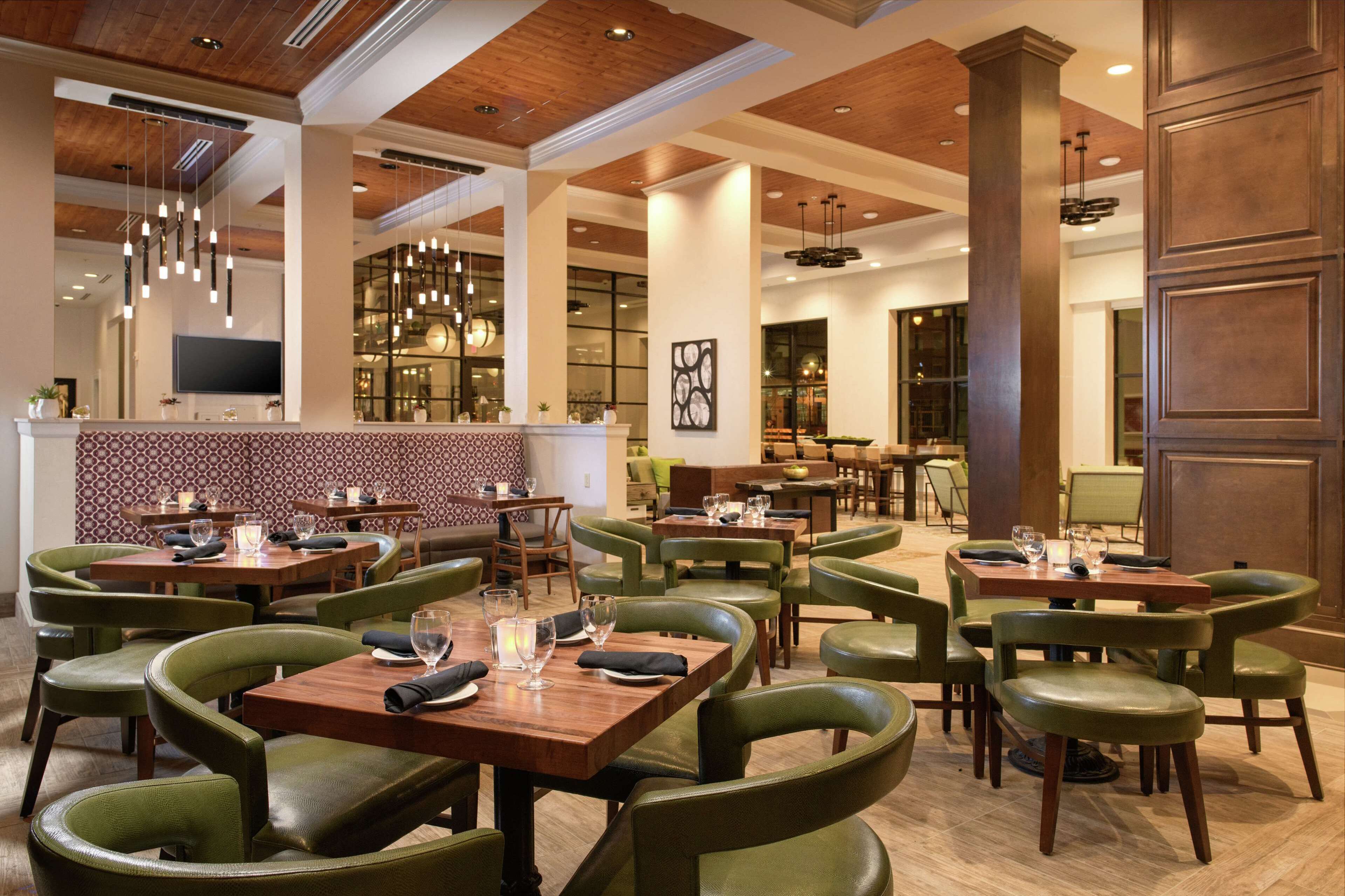 A spacious restaurant with green upholstered chairs, wooden tables, and hanging pendant lights.