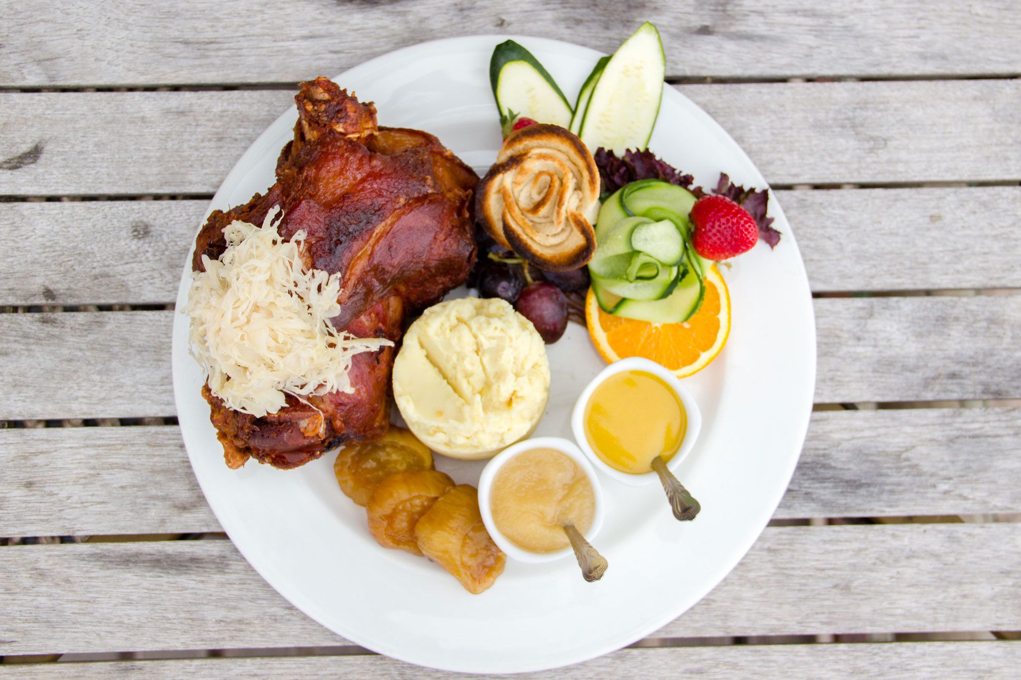 A plate with roasted meat, sauerkraut, mashed potatoes, pickles, and fruit on a wooden table.