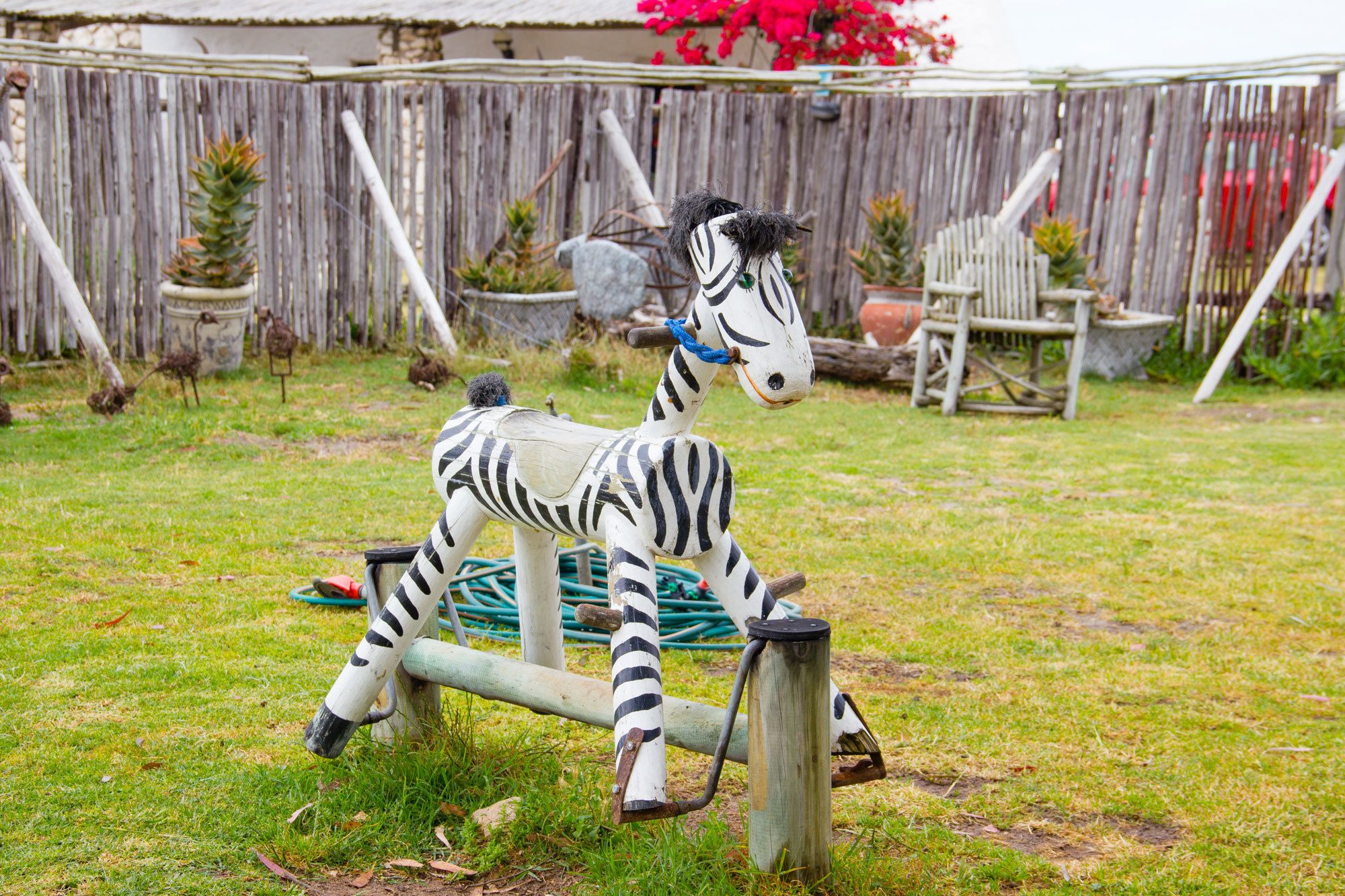 A zebra sculpture in a garden with a wooden fence, potted plants, and a chair.
