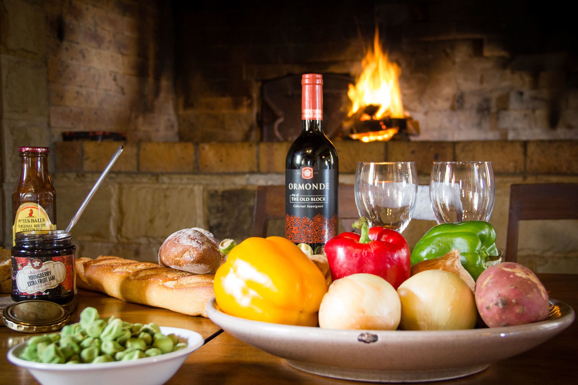 Una mesa con una botella de vino, dos copas, un cuenco de verduras y un frasco de mermelada.