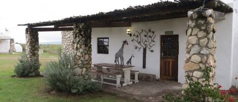 Un edificio rústico de piedra con una mesa y sillas de madera en el exterior, una escultura de jirafa y ciervo en la pared, y un pilar de piedra con una puerta.