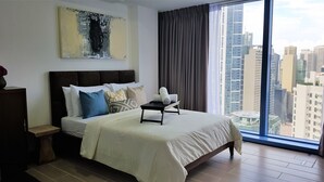 A modern hotel room with a large bed, a bedside table, a view of the cityscape through a large window, and a piece of abstract art on the wall. - Three Central (Makati)