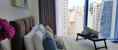 A modern hotel room with a large window, a bed with multiple pillows, a small table, and a vase of flowers.