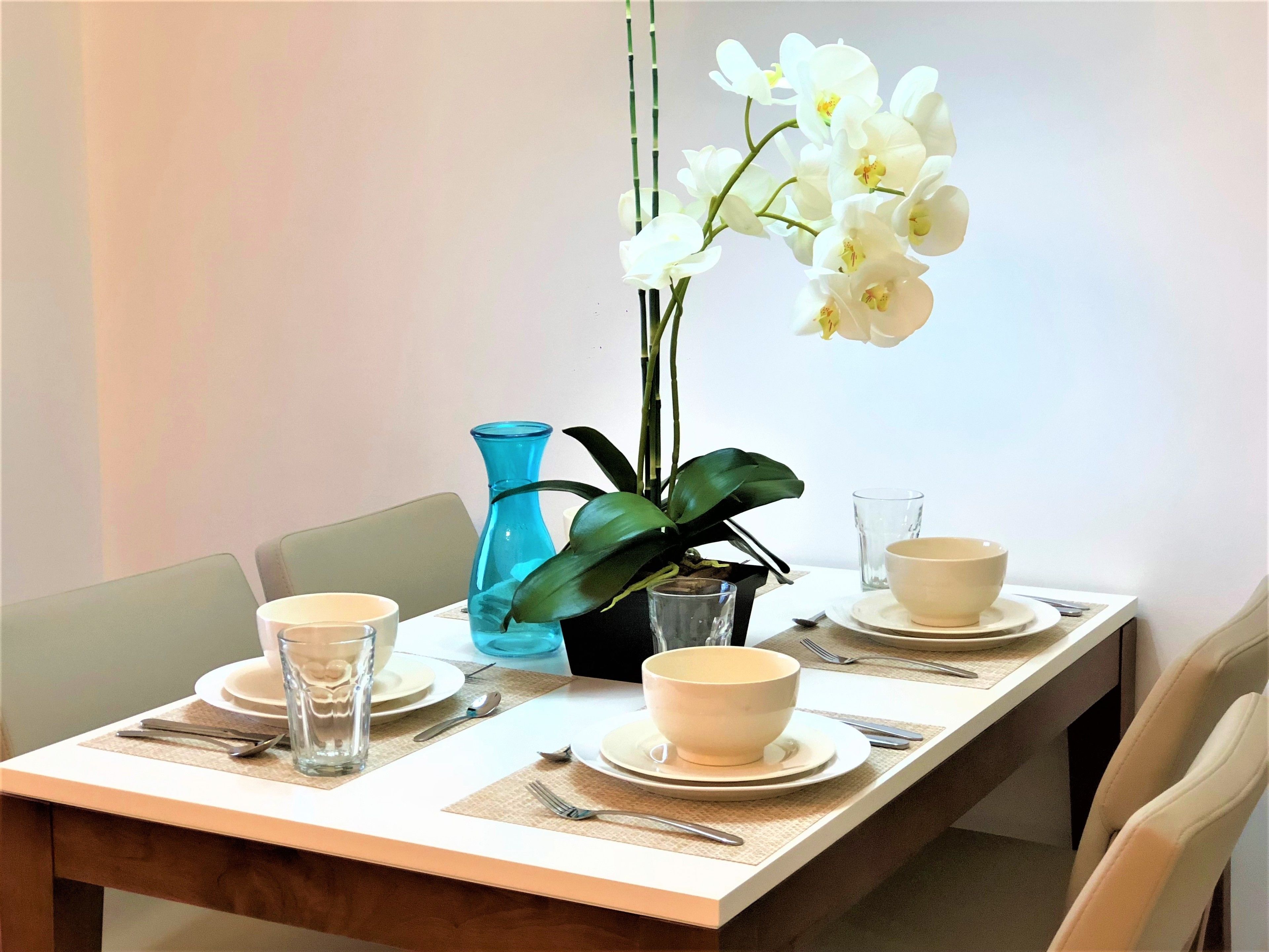 Una mesa de comedor dispuesta con platos, tazones, cubiertos y un florero con flores blancas.