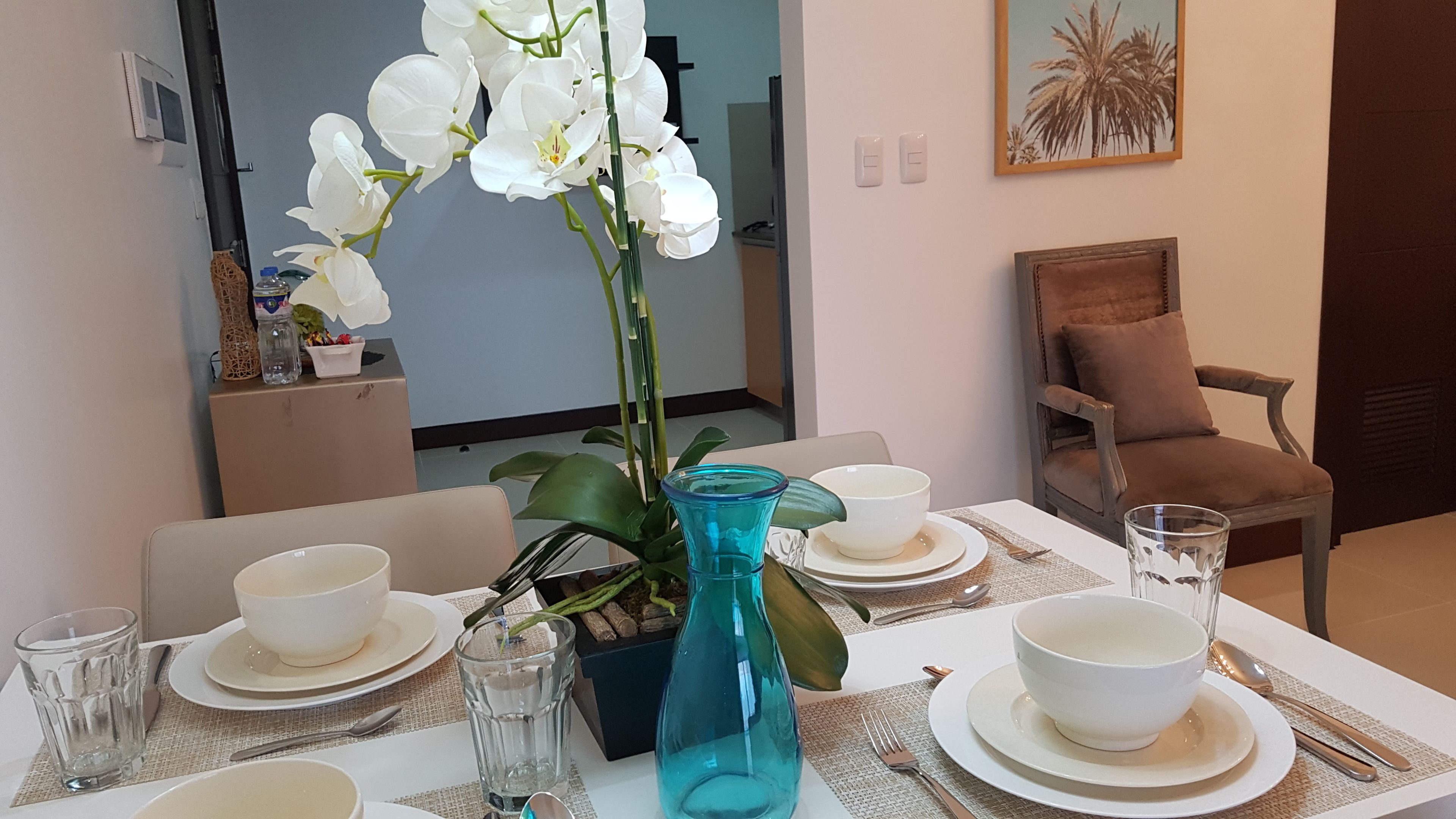 Une table à manger dressée avec de l’assiette, des bols et des couverts blancs, un vase bleu rempli de fleurs et une chaise agrémentée d’un coussin.