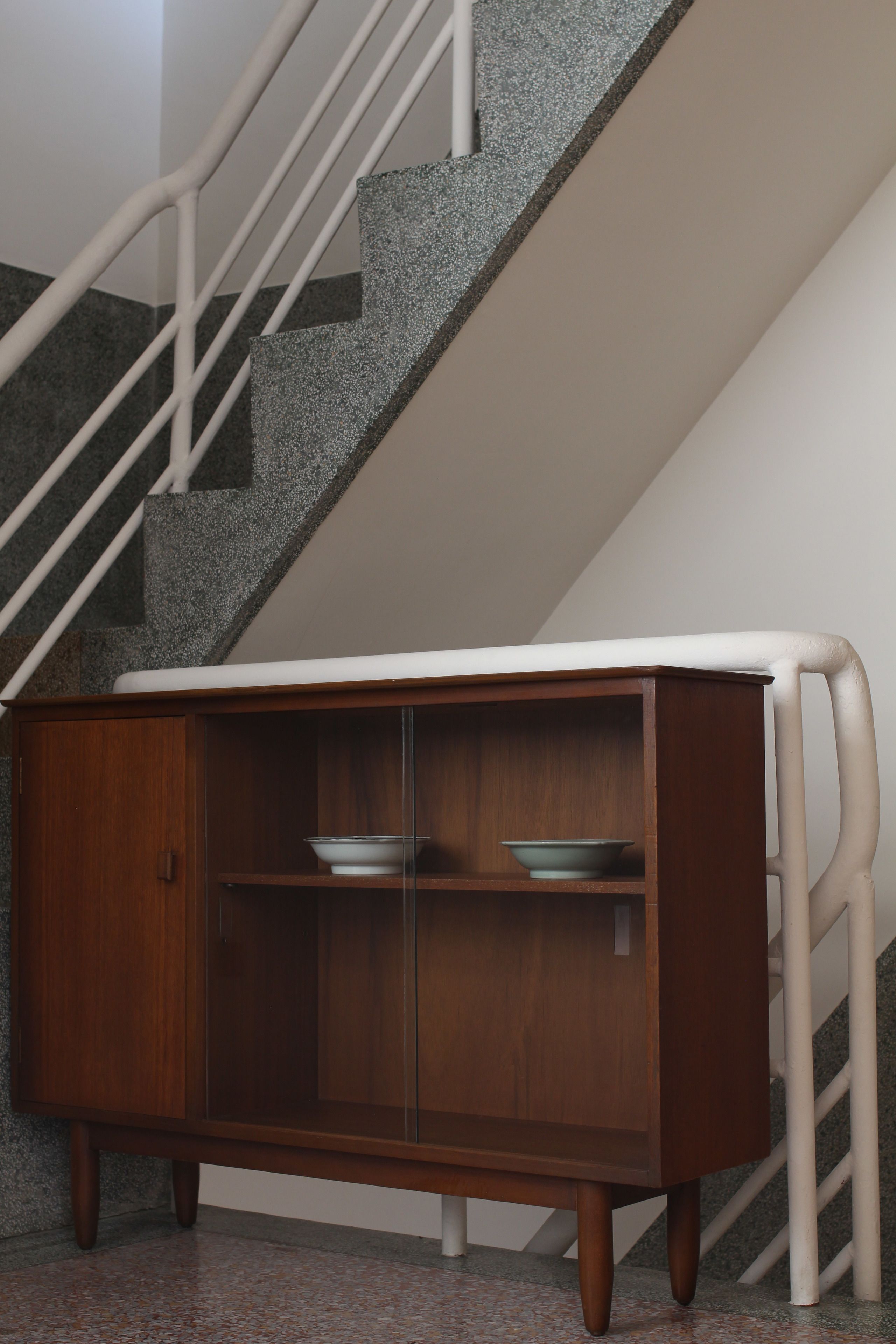 Un mueble de madera con puertas de cristal, situado frente a una escalera con barandillas blancas.
