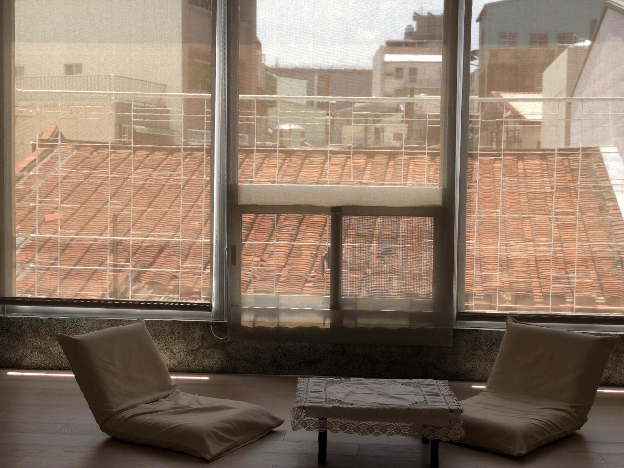 A room with two chairs and a small table, large windows with a view of a tiled roof and buildings.