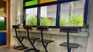 A room with a concrete floor, a window with a view of greenery outside, and a shelf with potted plants. - The Picturebook Guesthouse (Mae Sot)