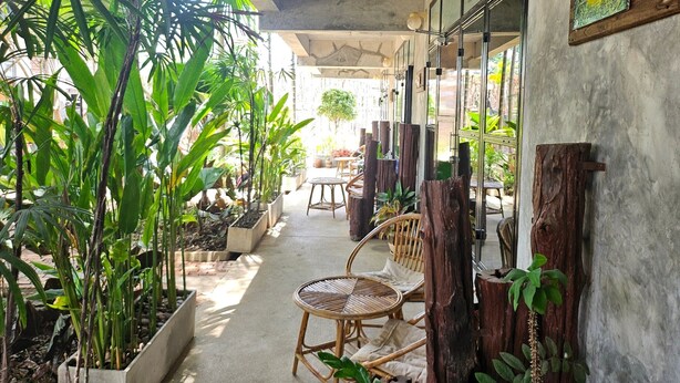 A covered outdoor area with potted plants, wicker furniture, and a concrete floor. - The Picturebook Guesthouse (Mae Sot)