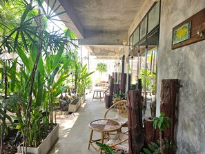 A covered outdoor area with potted plants, wicker furniture, and a concrete floor. - The Picturebook Guesthouse (Mae Sot)