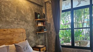 A bedroom with a wooden headboard, a bed with grey bedding, a nightstand, a shelf with books and a decorative item, and a window with a view of greenery. - The Picturebook Guesthouse (Mae Sot)