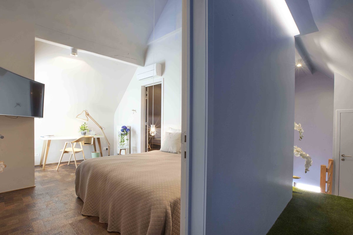 A bedroom with a bed, a desk, a chair, a TV, and a door leading to another room.