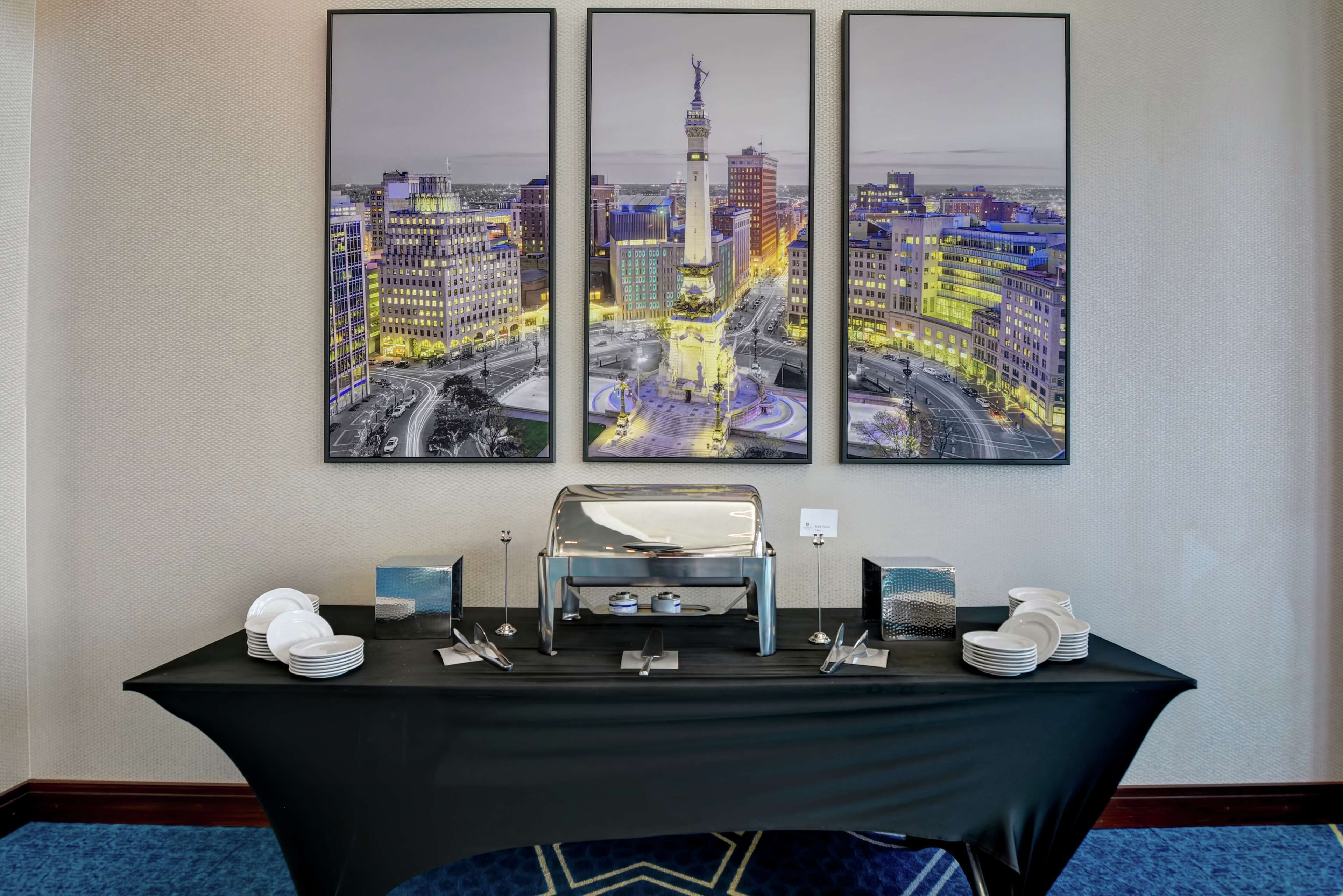 Een buffettafel met een zwarte tafelkleed, witte borden en een zilveren buffetstelling, voor een muur met drie kunstwerken van stadsgezichten.