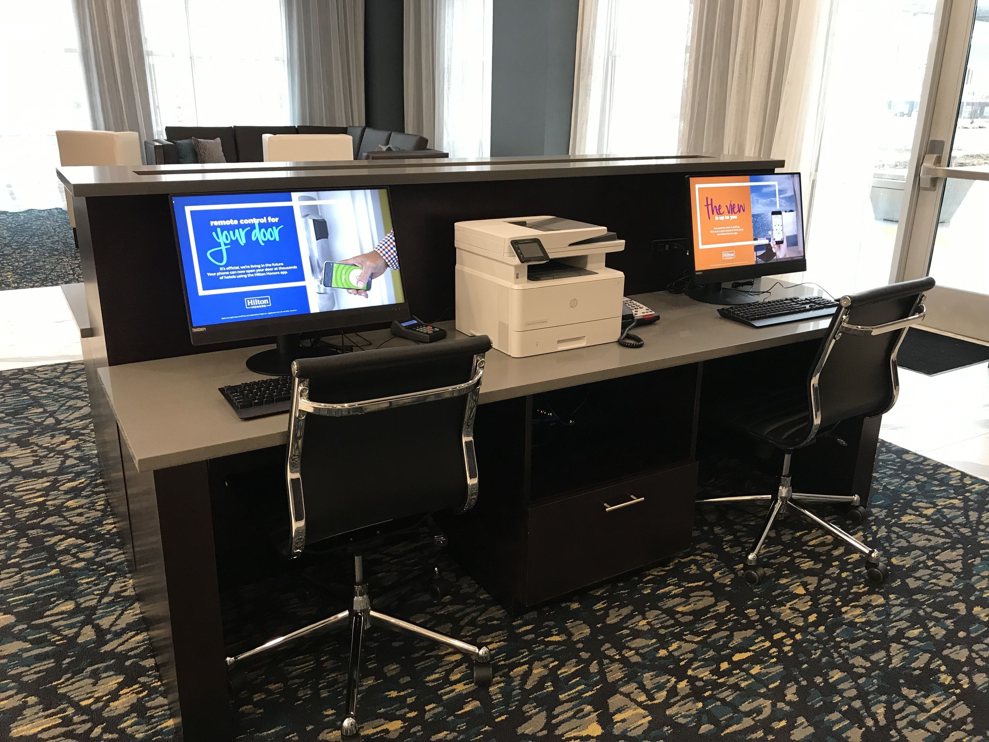 A hotel reception desk with two monitors, a printer, a telephone, and two chairs.