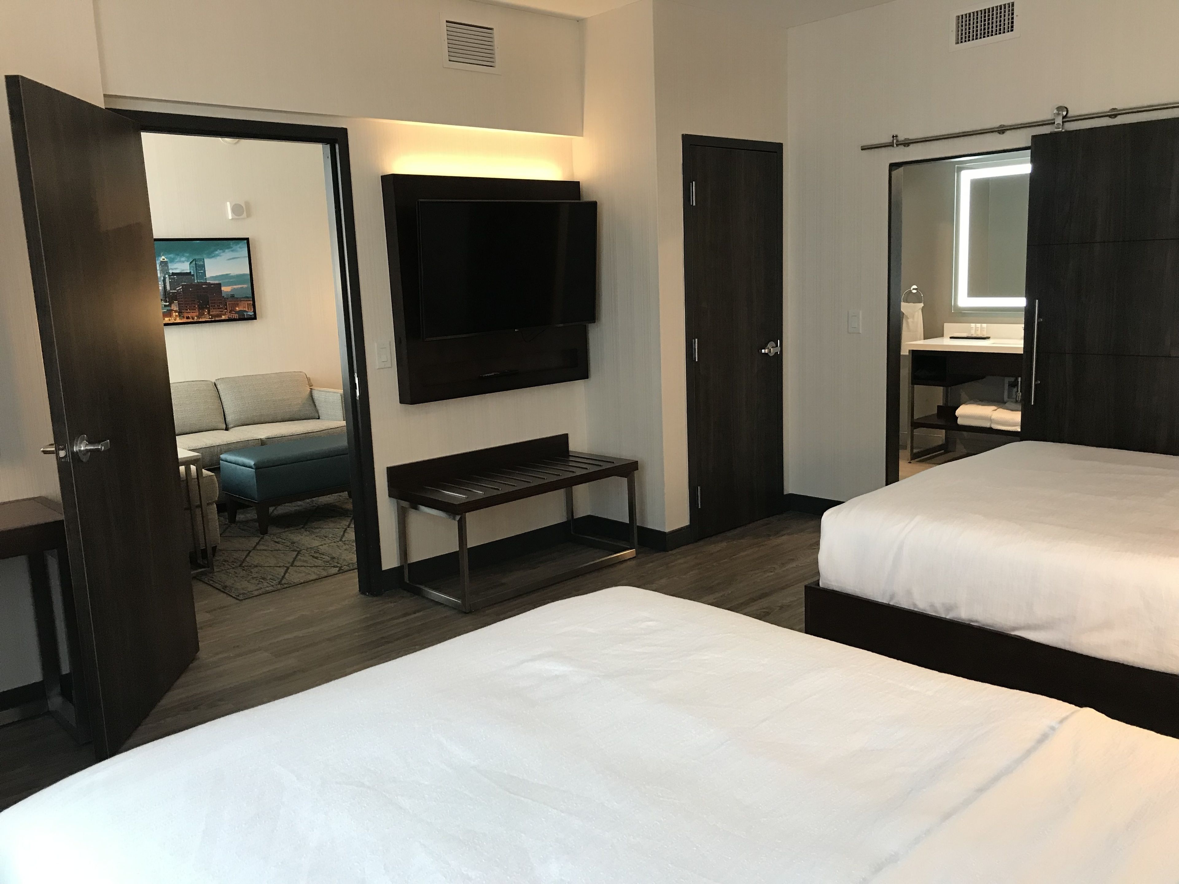 A hotel room with two beds, a TV mounted on the wall, a bench, and a door leading to a bathroom.