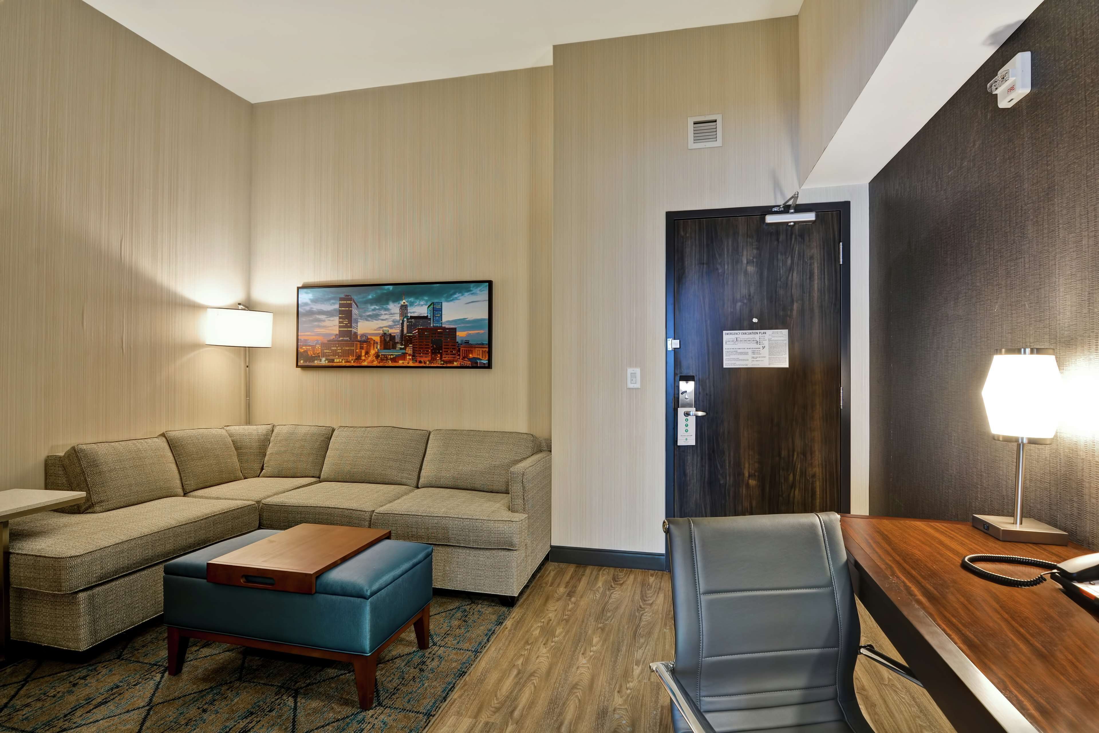 A hotel room with a sectional sofa, a small table, a desk with a chair, a lamp, and a painting on the wall.