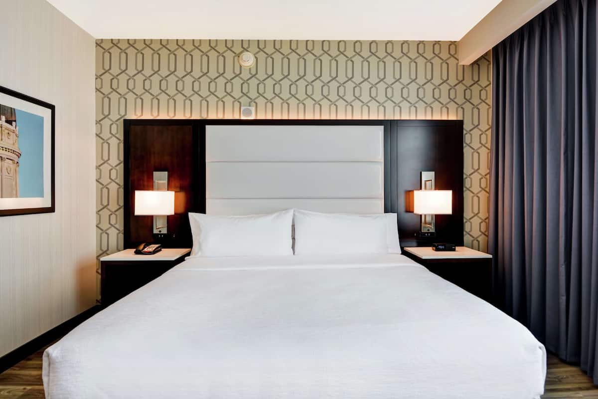 Une chambre d’hôtel avec un grand lit, deux tables de chevet avec des lampes, un papier peint à motifs et un tableau encadré au mur.