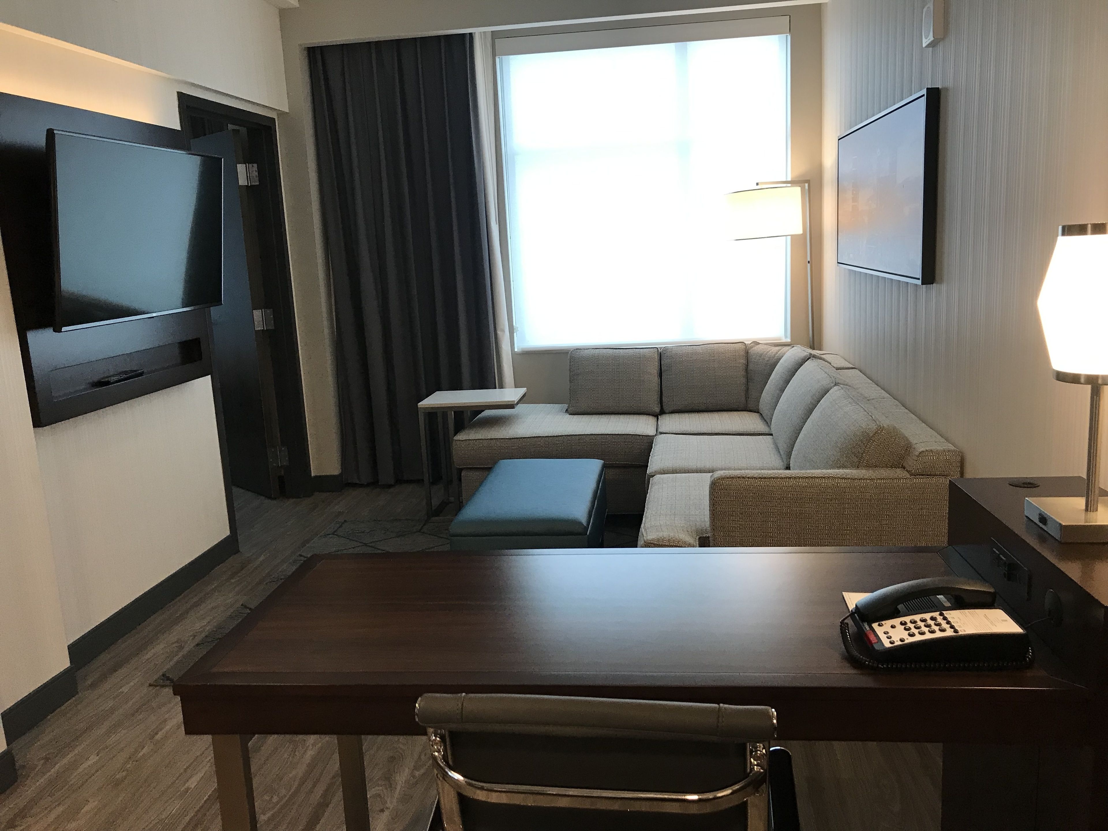 Een hotelkamer met een flatscreen televisie aan de muur, een bank, een klein tafeltje, een bureau met stoel en een bedlampje.