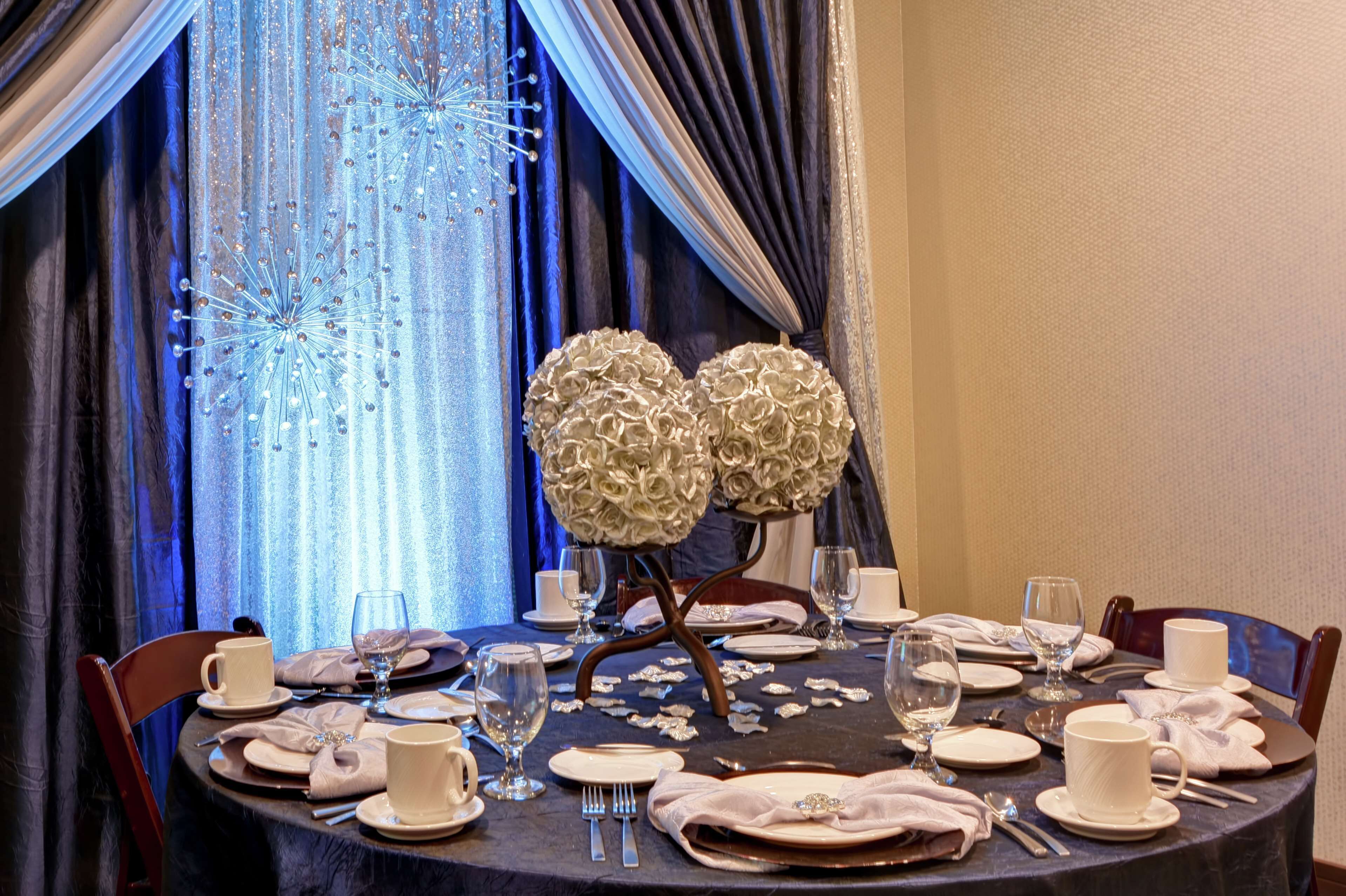 A dining table set for a meal with a decorative centerpiece, elegant tableware, and a view of a blue curtain with a sparkling effect.