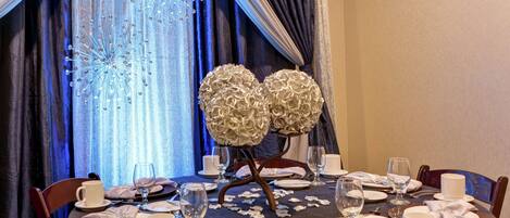 A dining table set for a meal with a decorative centerpiece, elegant tableware, and a view of a blue curtain with a sparkling effect.