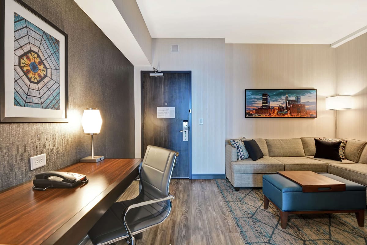 Une chambre d’hôtel avec un bureau en bois, une chaise, un canapé et un tableau représentant un paysage urbain.