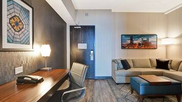 A hotel room with a wooden desk, a chair, a sofa, and a cityscape painting.