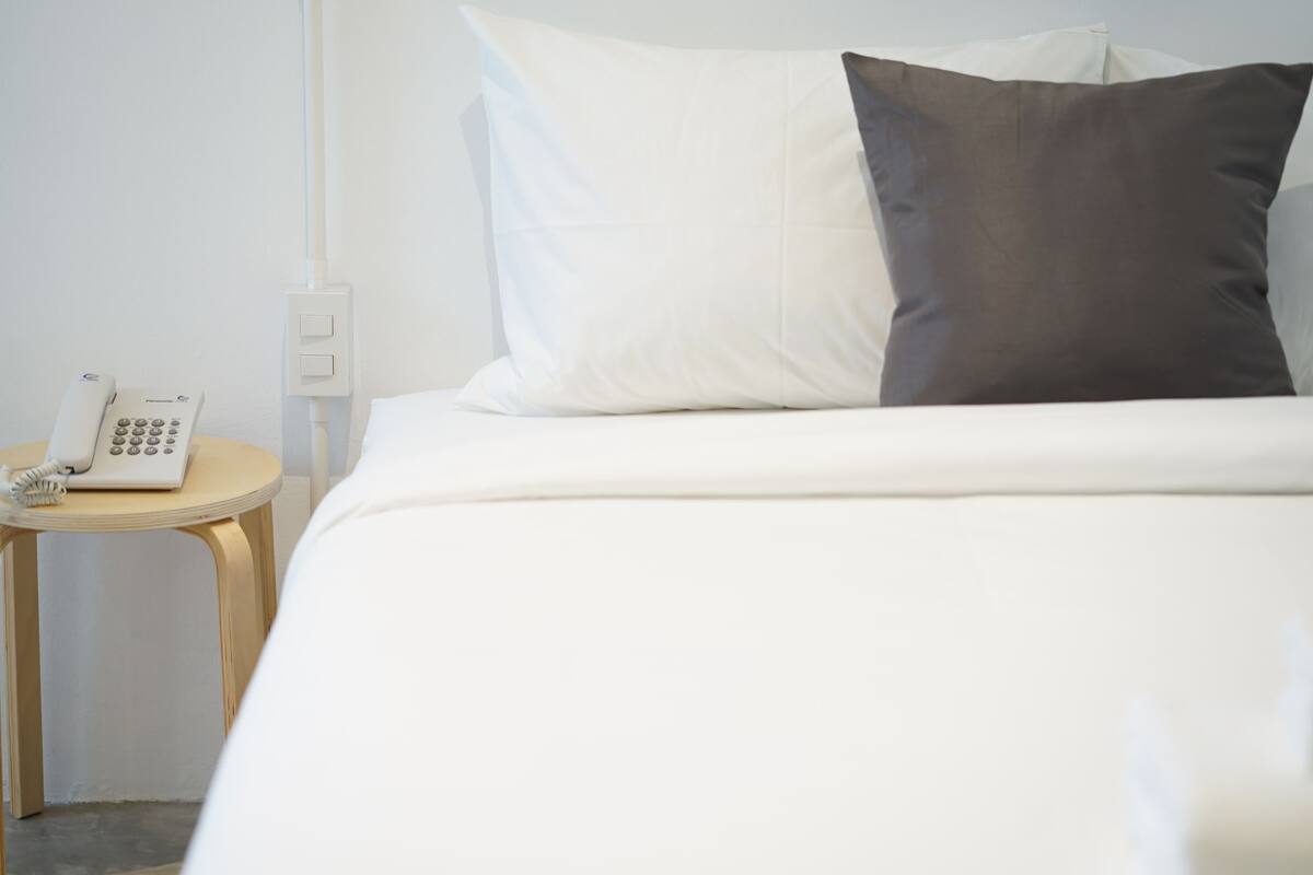 A hotel room with a bed, white and dark gray pillows, a bedside table with a phone, and a wall socket.