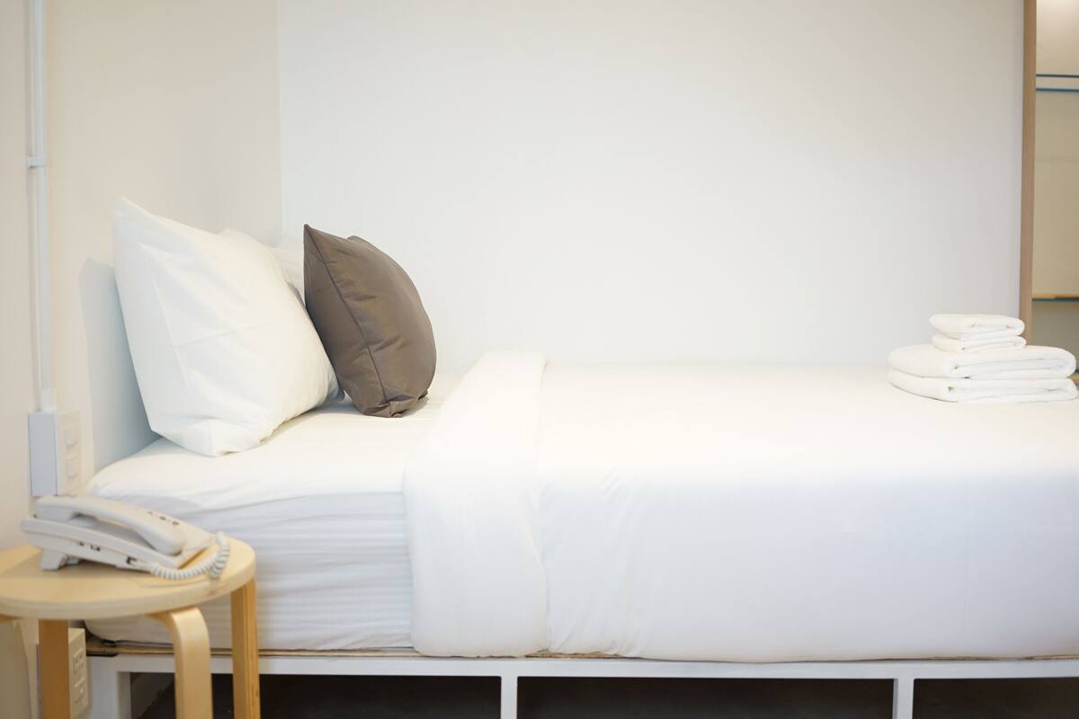 A neatly made bed with white and brown pillows, a white blanket, and a stack of white towels on the right side.