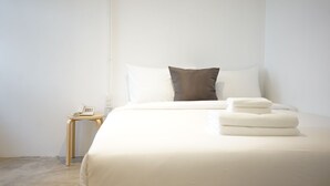 A neatly made bed with white sheets and two pillows, a small bedside table with a phone, and a light-colored floor.