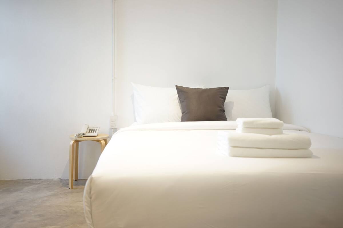 A neatly made bed with white sheets and two pillows, a small bedside table with a phone, and a light-colored floor.