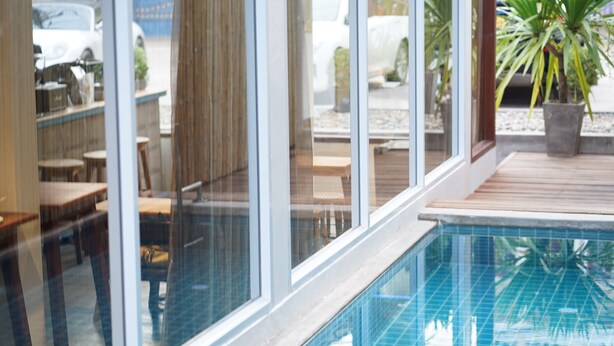 An indoor swimming pool with clear glass walls, wooden deck, and potted plants.