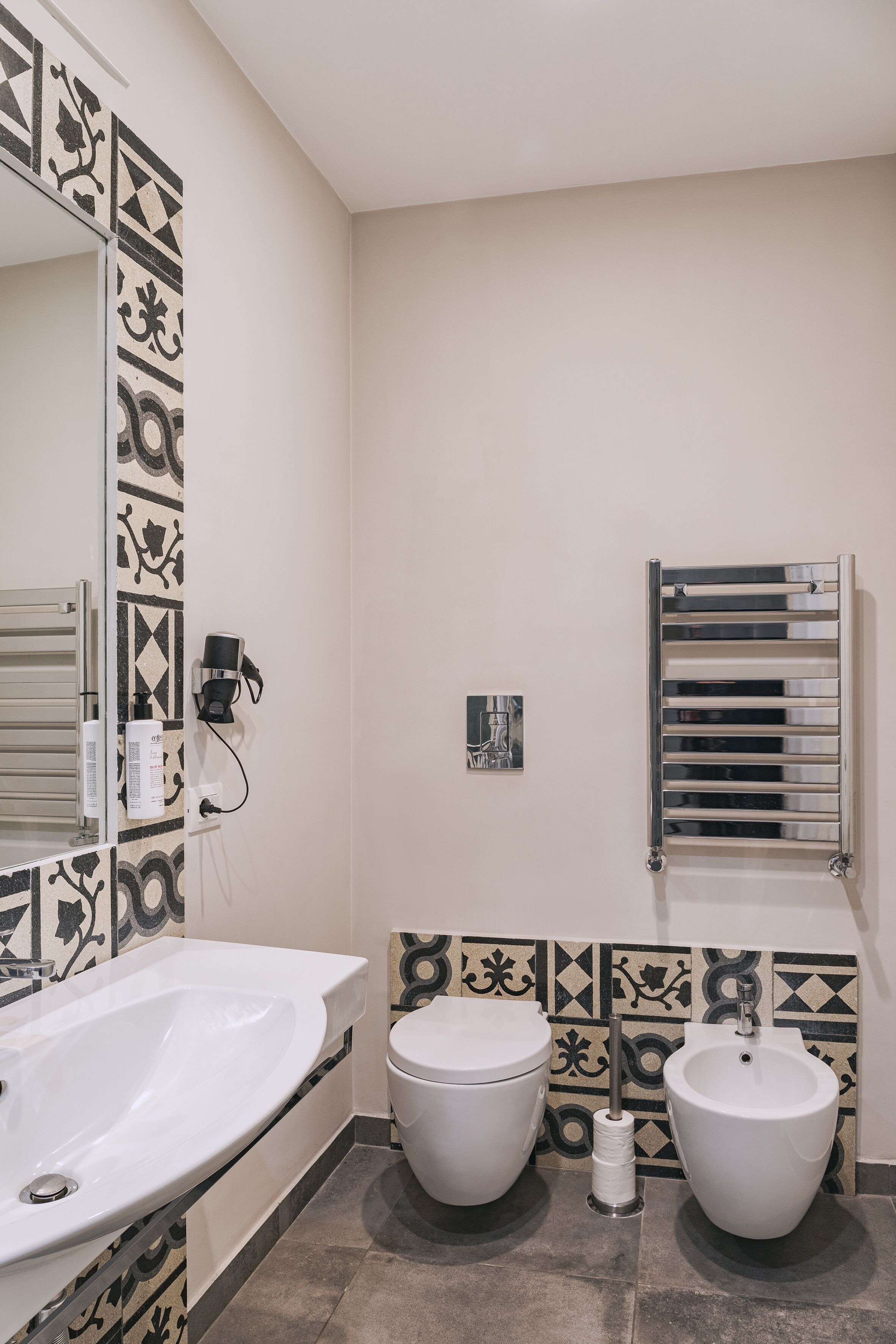 A bathroom with a white sink, two toilets, a towel rack, and patterned tiles on the walls.