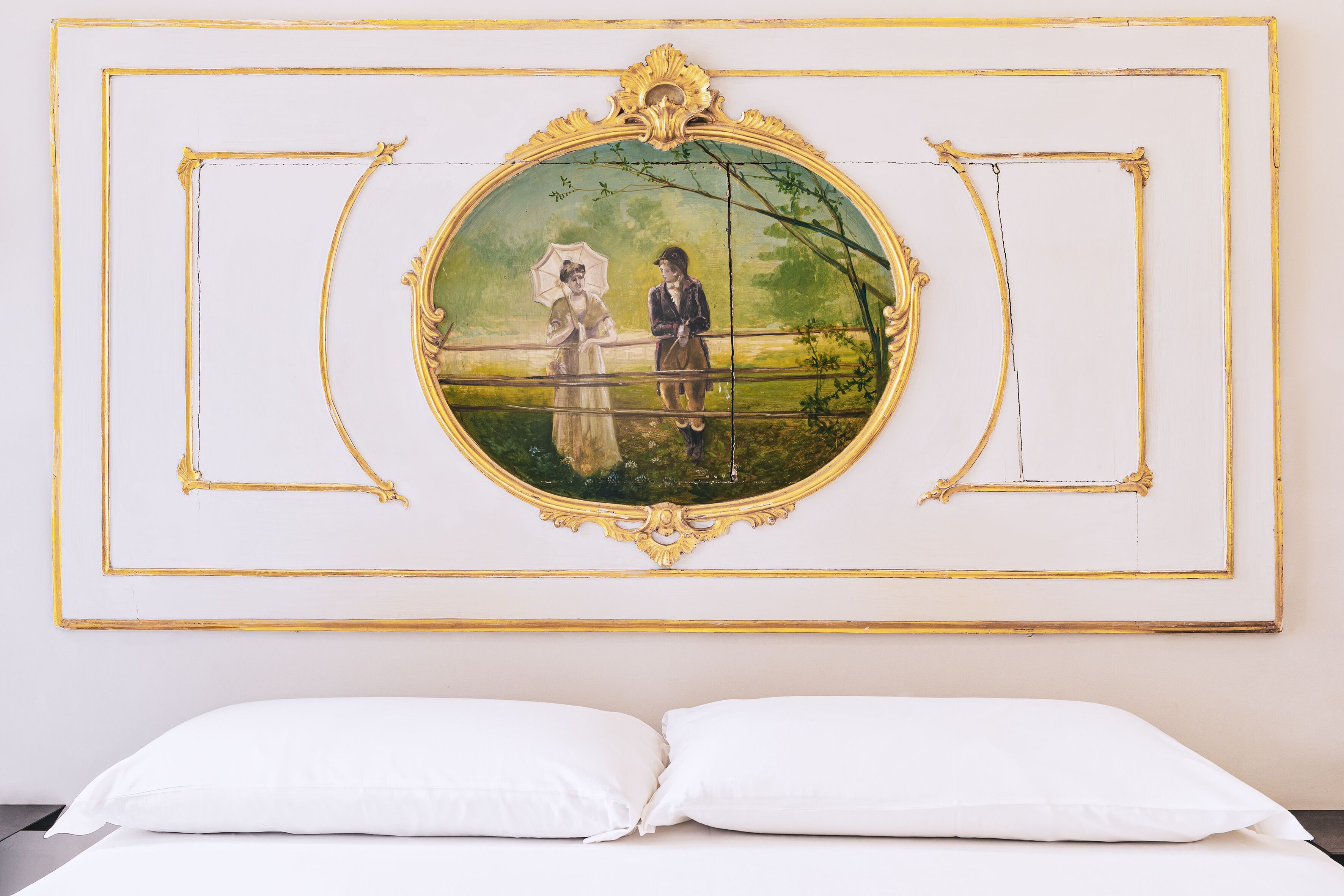 A room with a bed, white pillows, and a large framed painting of a couple on a bridge.