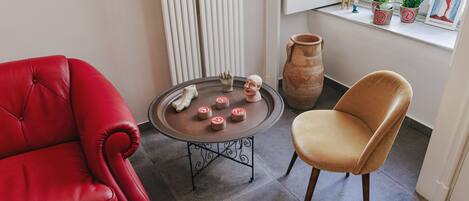 A room with a red leather sofa, a brown leather chair, a small round table with decorative items, a large clay pot, and a window with potted plants.