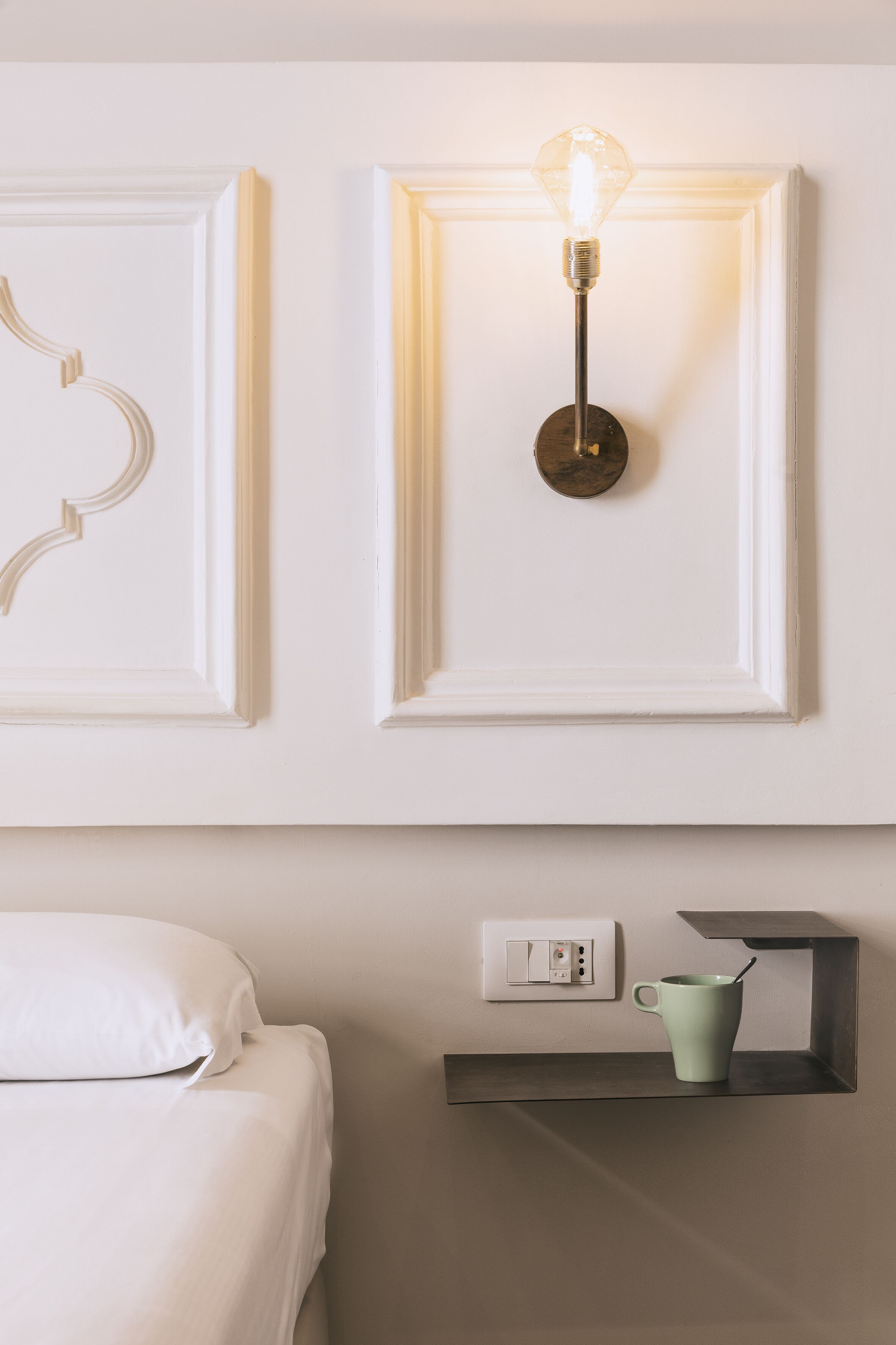 A minimalist hotel room with a bed, a wall-mounted light fixture, and a small shelf with a mug.