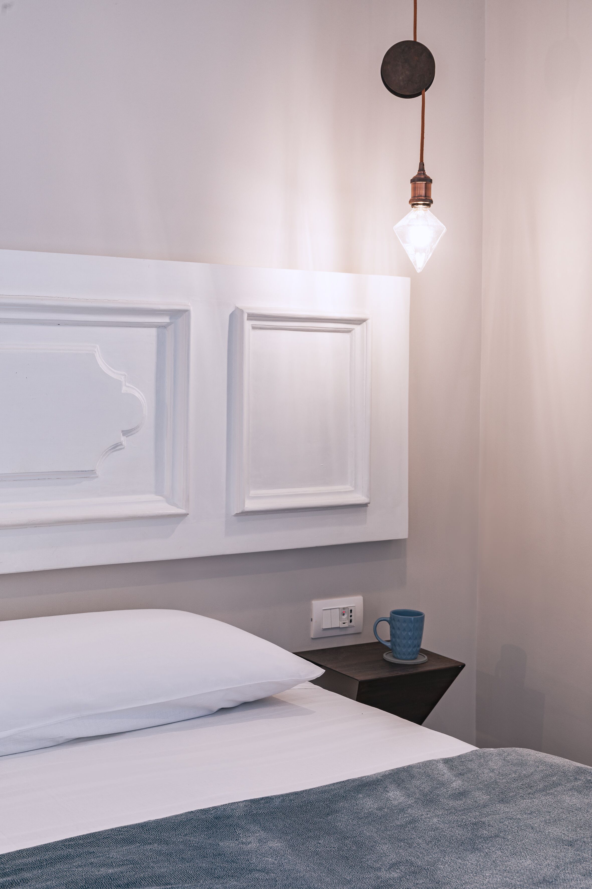 Una cama bien hecha con un almohadón blanco y una manta azul, una mesita de noche con una taza azul, y una lámpara de pared.