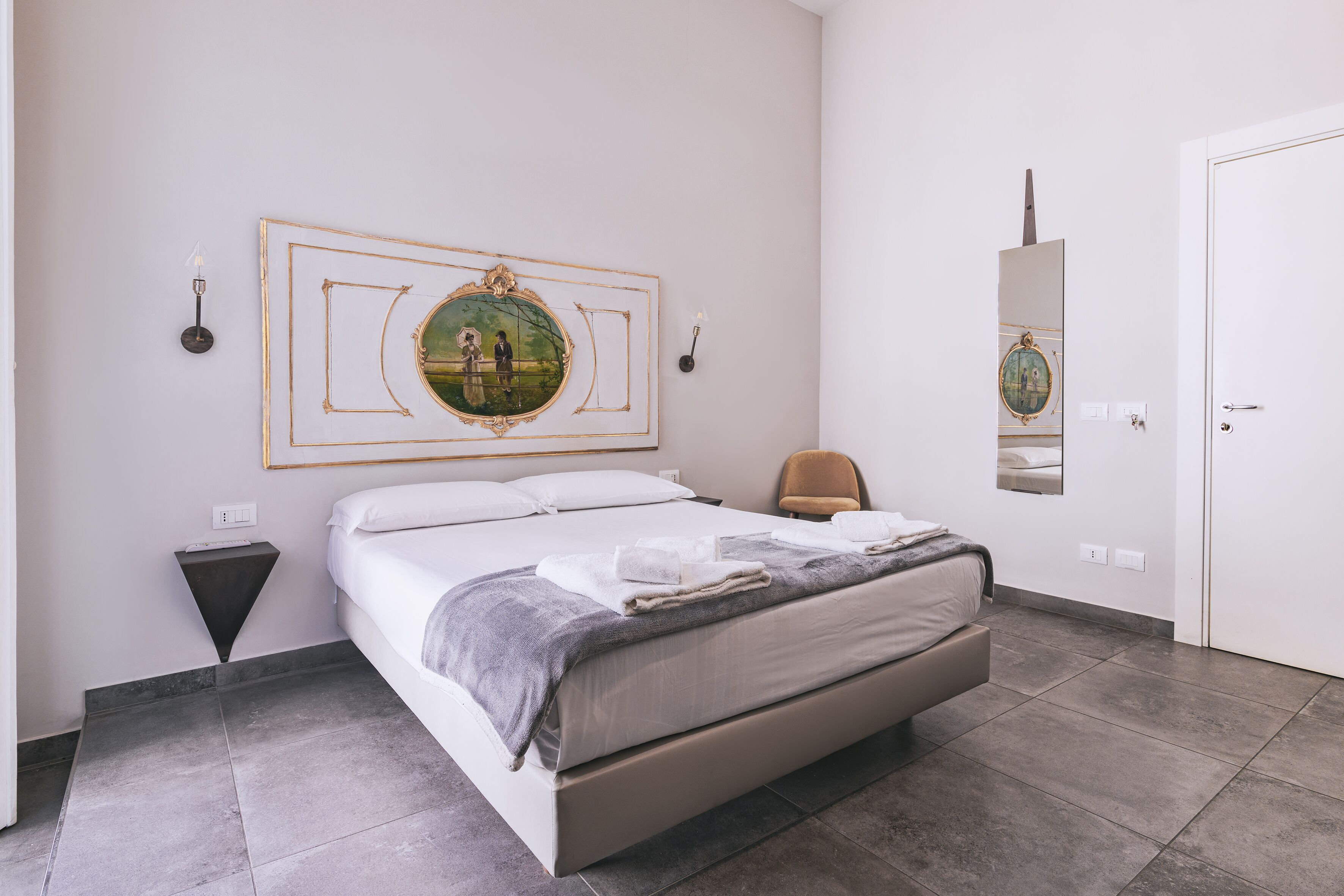 Ein ordentlich eingerichtetes Schlafzimmer mit einem großen Bett, einem Nachttisch, einem Sessel, einem Wandspiegel und einem gerahmten Bild über dem Bett.