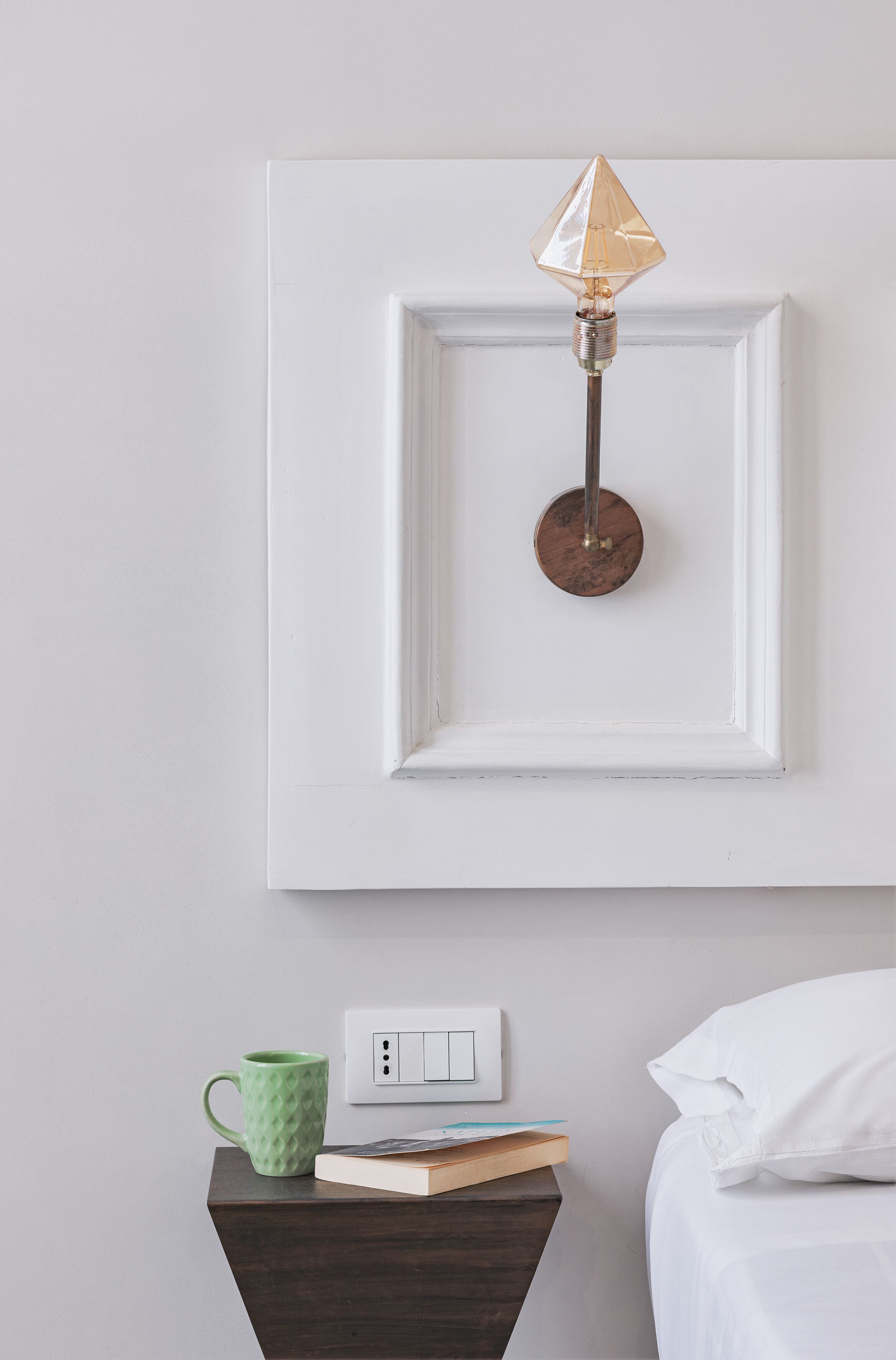 Une pièce bien rangée, avec un tableau encadré au mur, une table de chevet sur laquelle se trouvent une tasse verte, un livre et un oreiller blanc.