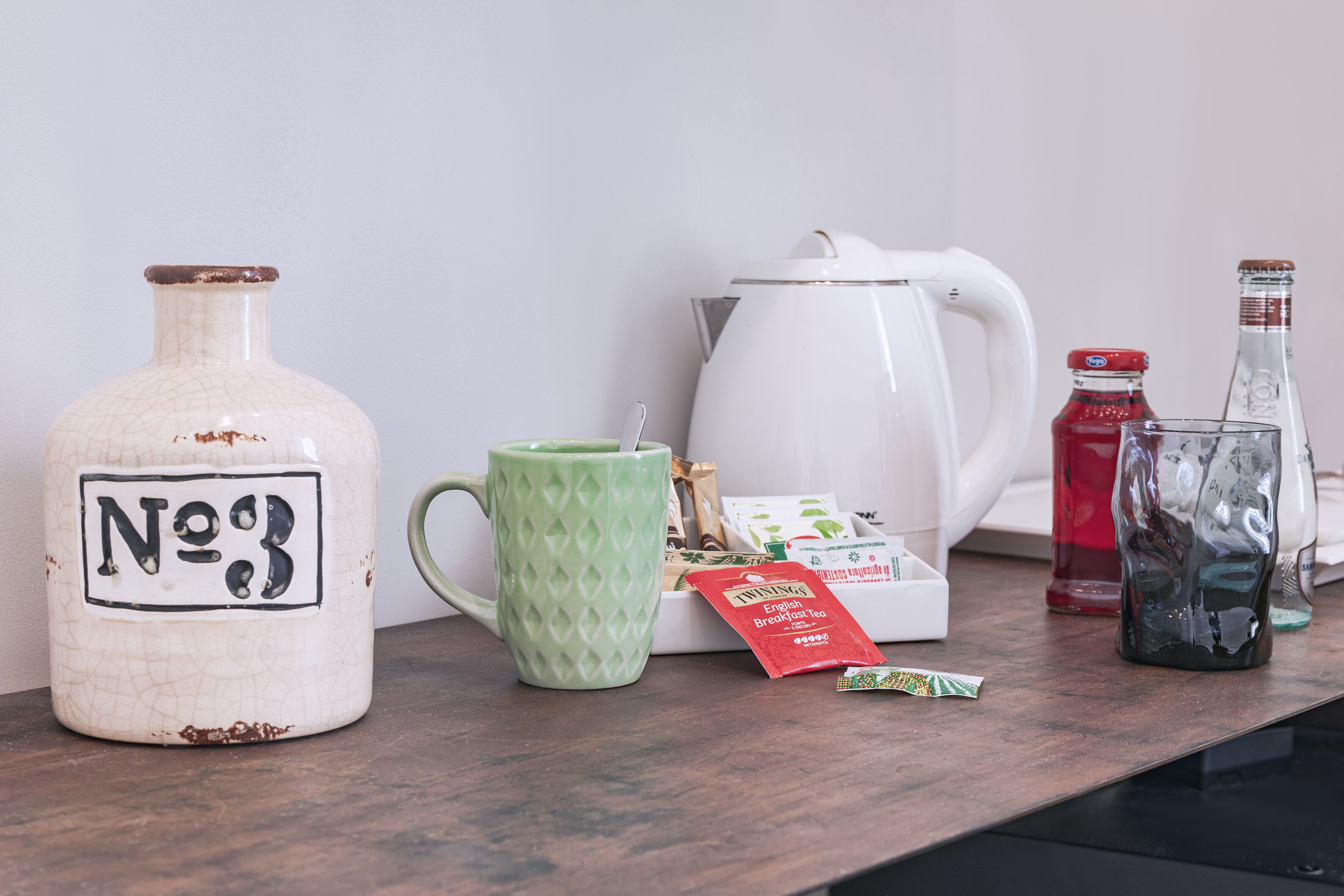 A kitchen counter with a white teapot, a green mug, a white pitcher, a red bottle, a clear bottle, and a packet of tea.