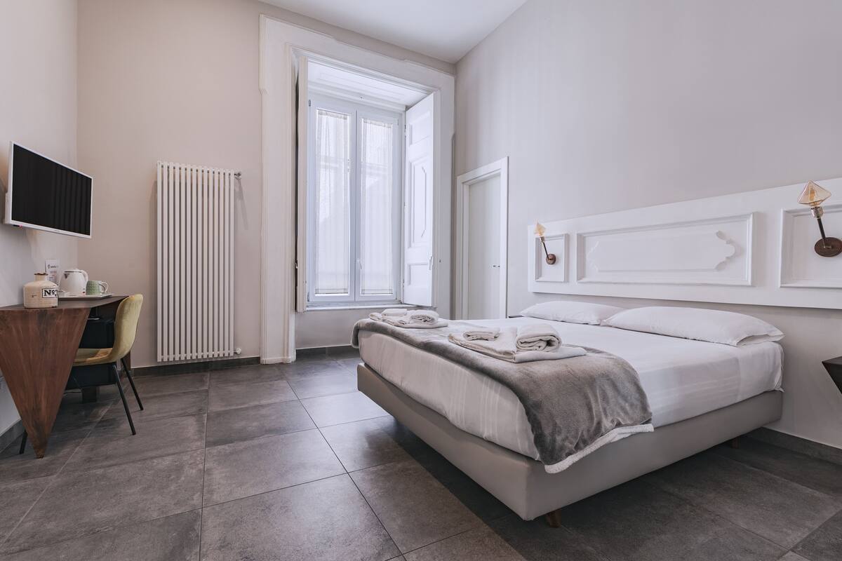 A modern hotel room with a bed, a desk, a chair, a radiator, and a window.