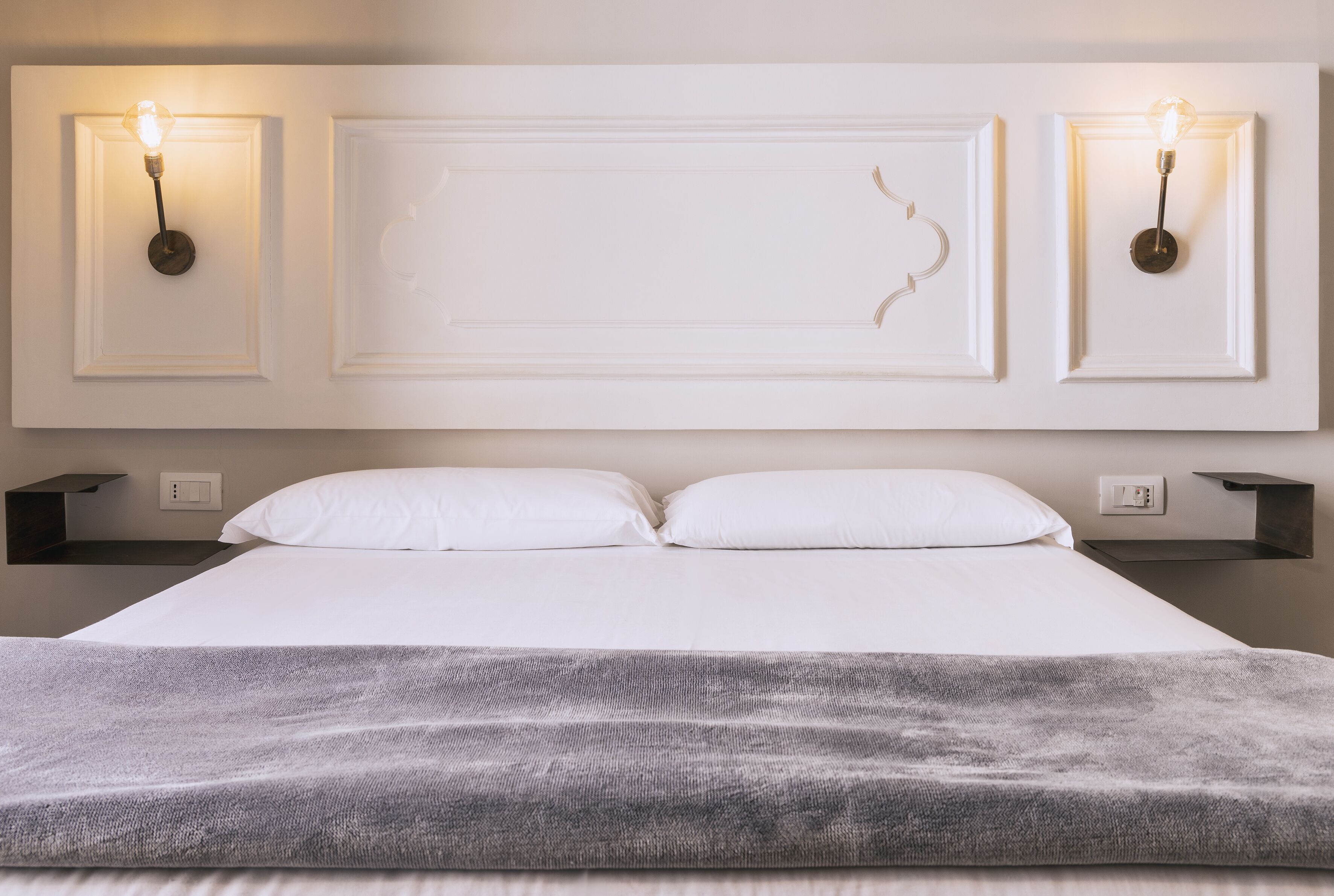Una cama bien hecha con sábanas blancas, un plaid gris y dos lámparas de pared con bombillas a la vista.