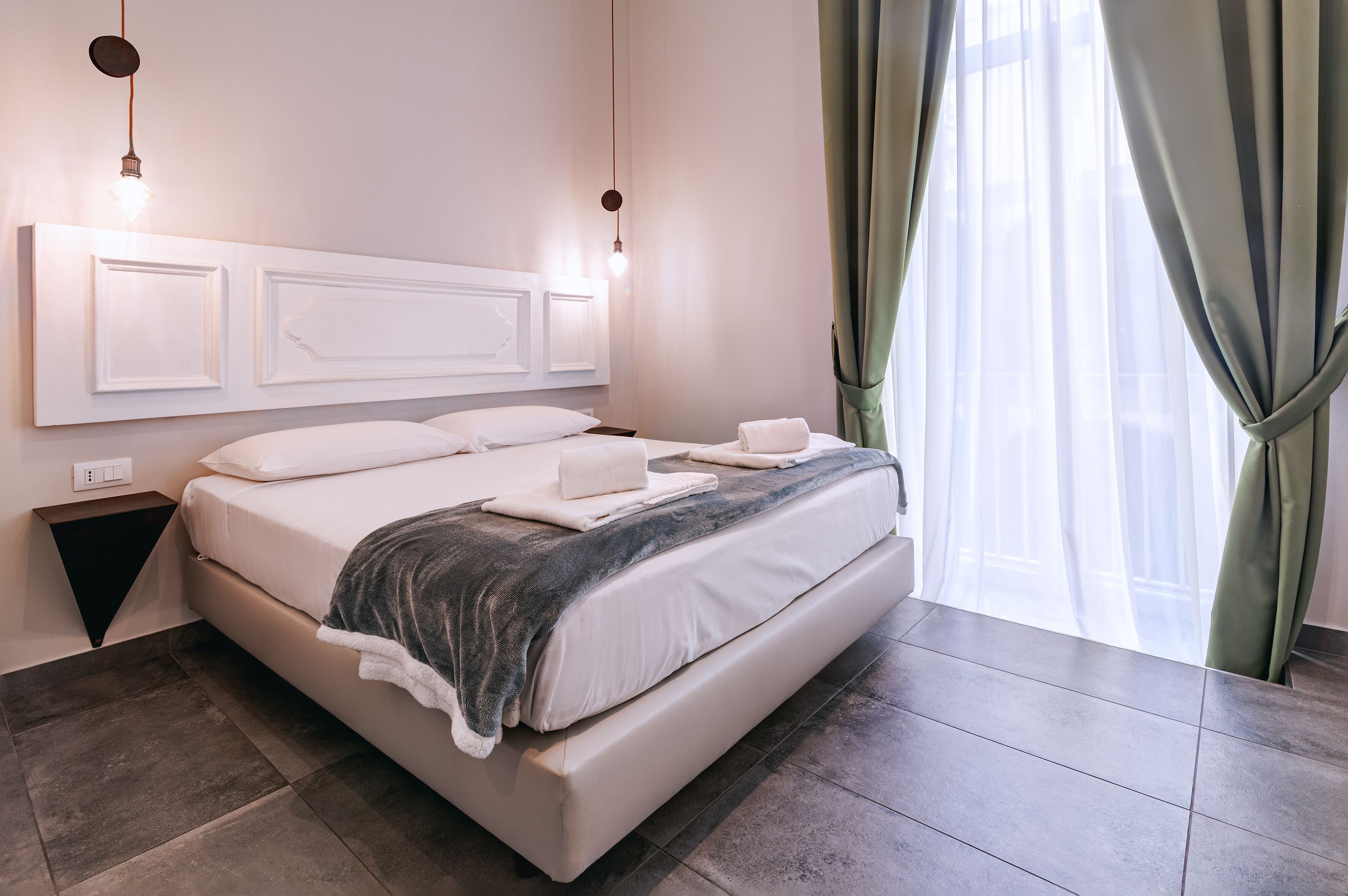A neatly made bed with white linens and a gray blanket, a bedside table with a lamp, and a view of a building through sheer curtains.